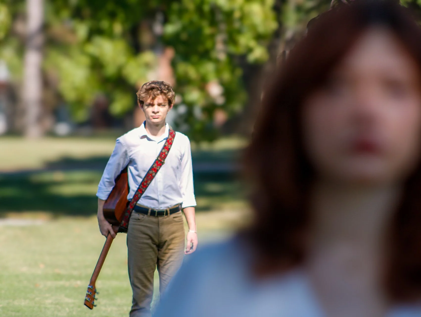 Young man carrying an acoustic guitar across a sunny park, viewed past a blurred person in the foreground.