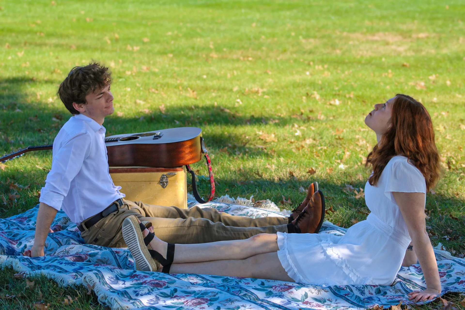 Young couple relaxing on a floral picnic blanket in a park beside an acoustic guitar and instrument case