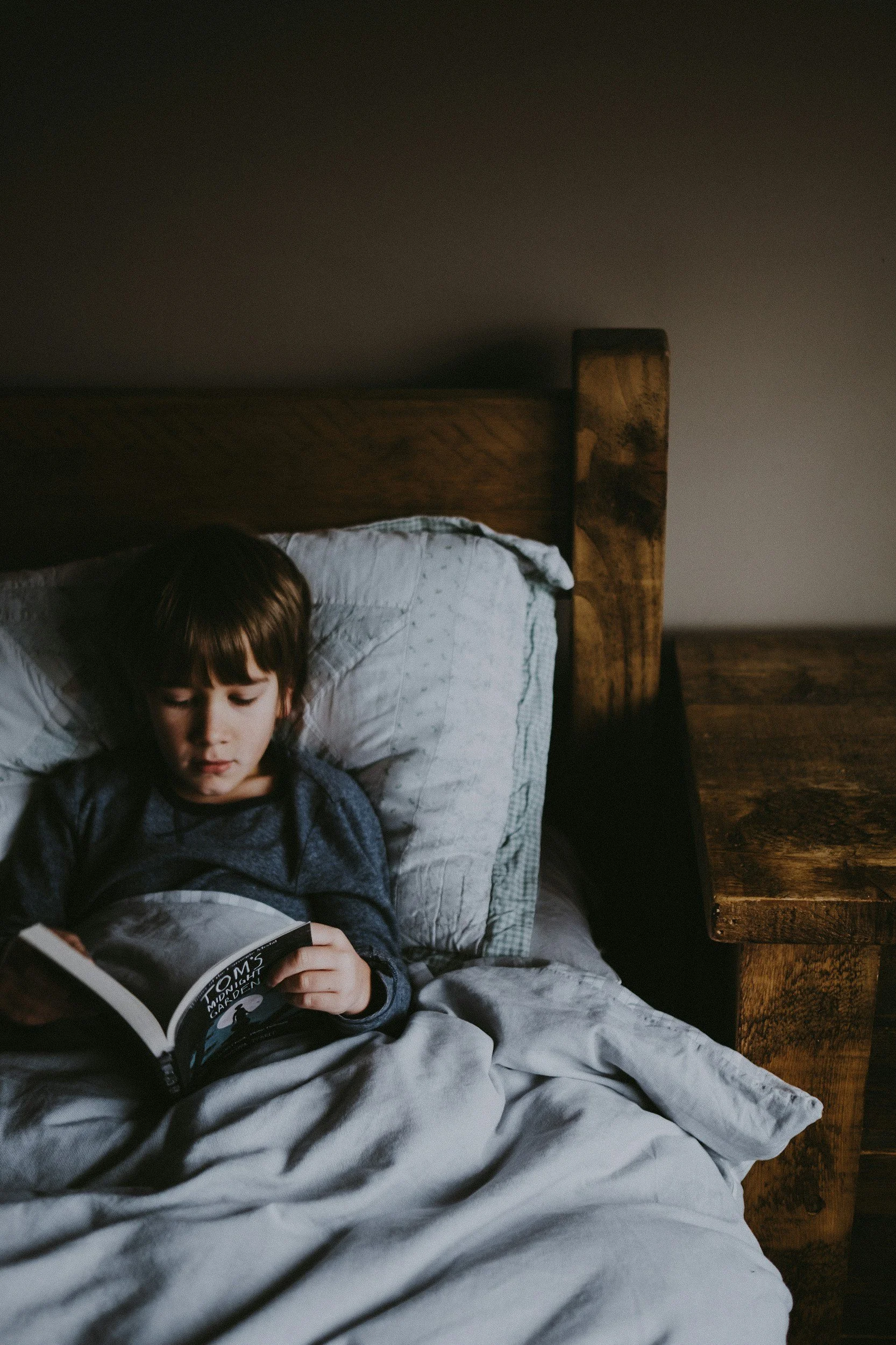 Enfant lisant un livre au lit, sous une couverture grise, dans une chambre chaleureuse en bois.