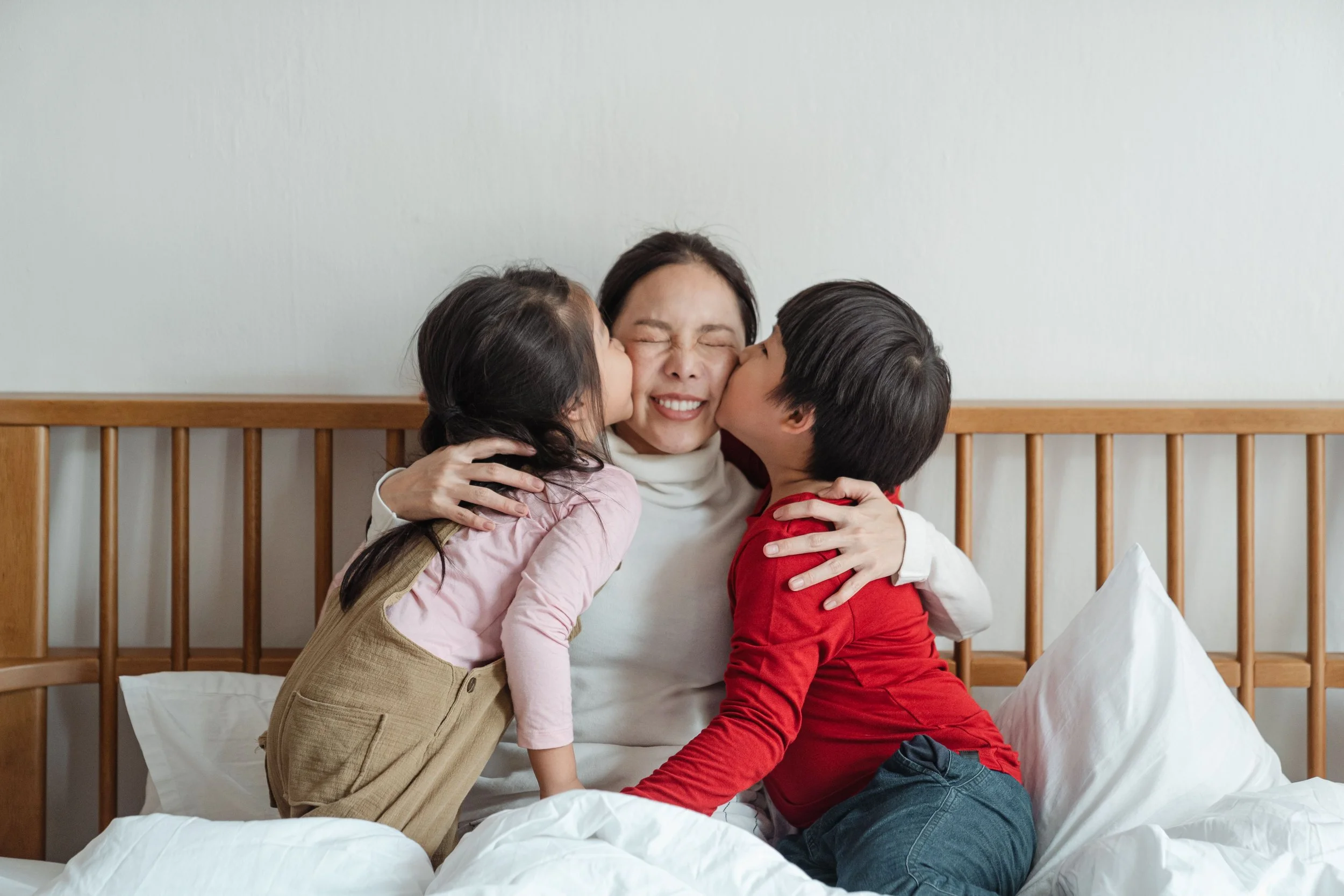 Une mère souriante serre ses deux enfants dans ses bras sur un lit, tandis qu’ils l’embrassent sur les joues.