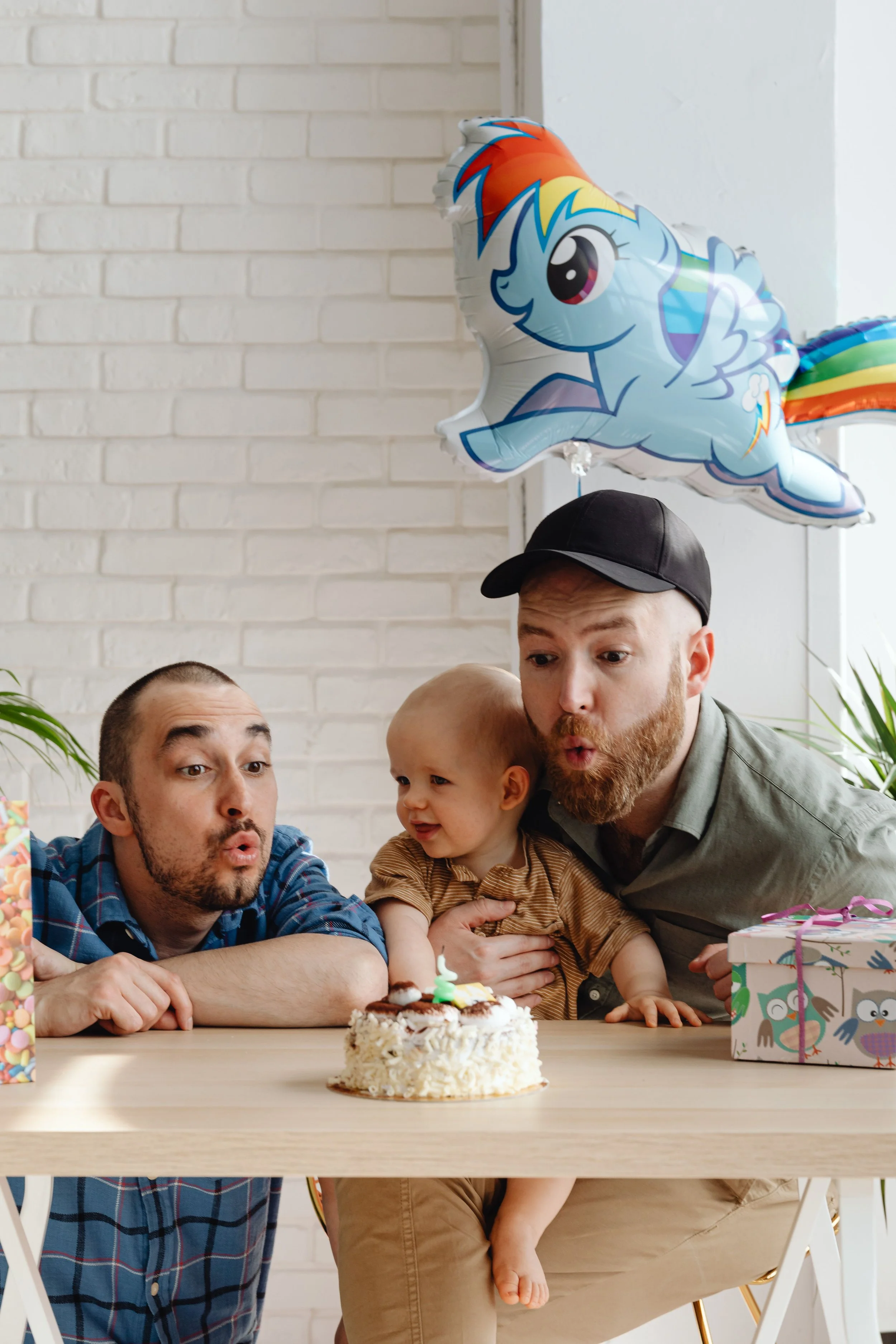 Deux hommes fêtent un anniversaire avec un bébé autour d’un gâteau, entourés de cadeaux et d’un ballon arc-en-ciel.