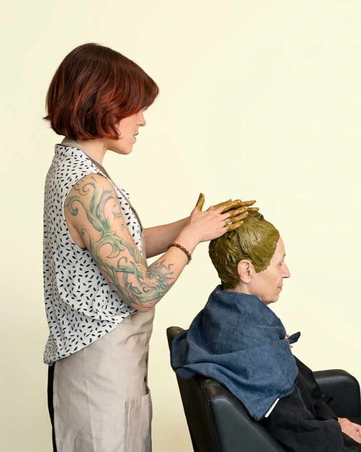 Une femme à la peau claire et aux cheveux roux applique un masque coloré vert foncé sur la tête d'une autre femme assise, qui porte une cape de salon de coiffure, dans un salon.