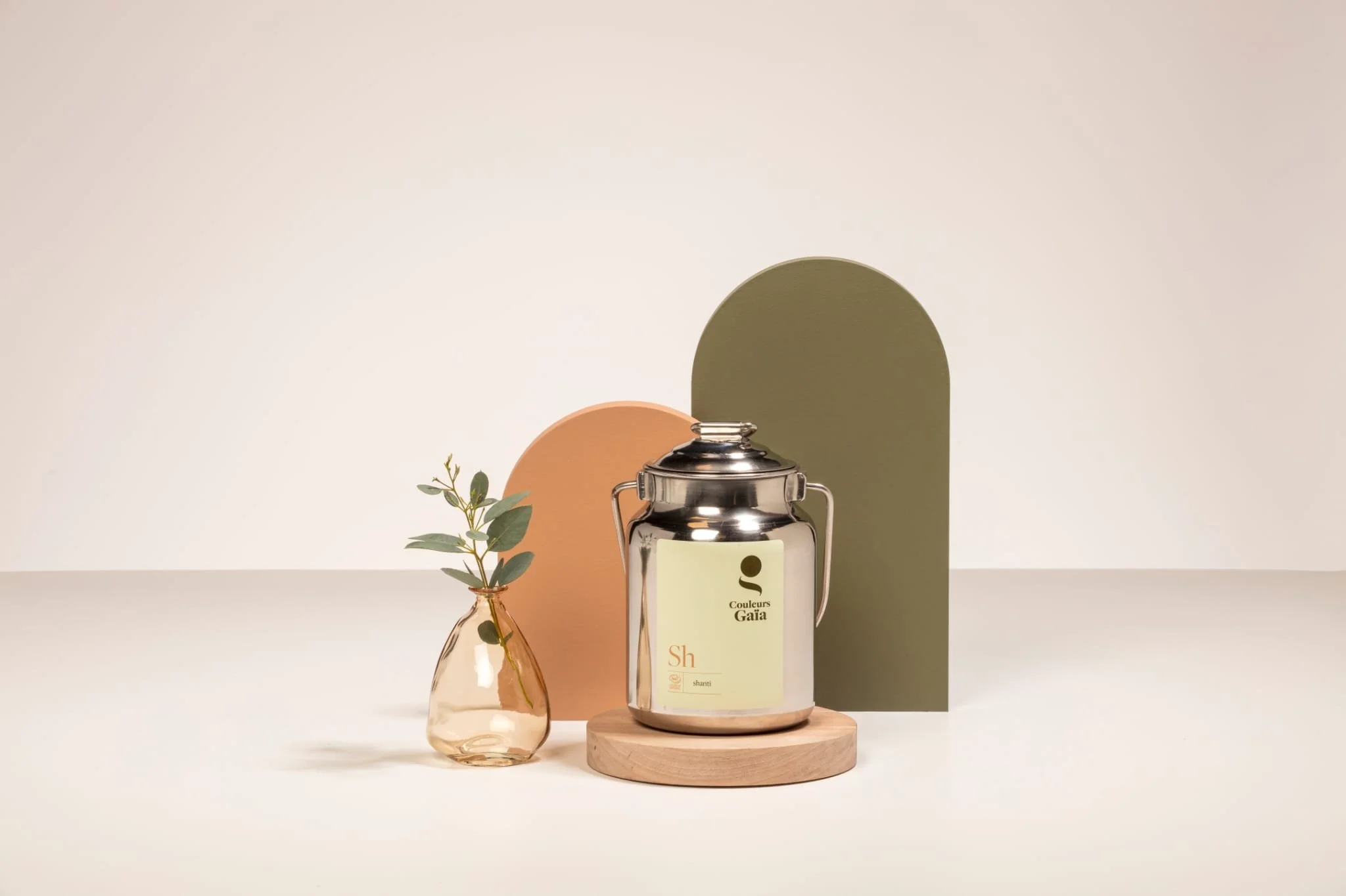 Une bouteille de parfum claire avec une étiquette jaune, deux panneaux décoratifs marron et vert derrière, une petite branche avec des feuilles dans un petit vase en verre, le tout posé sur une plateforme en bois.