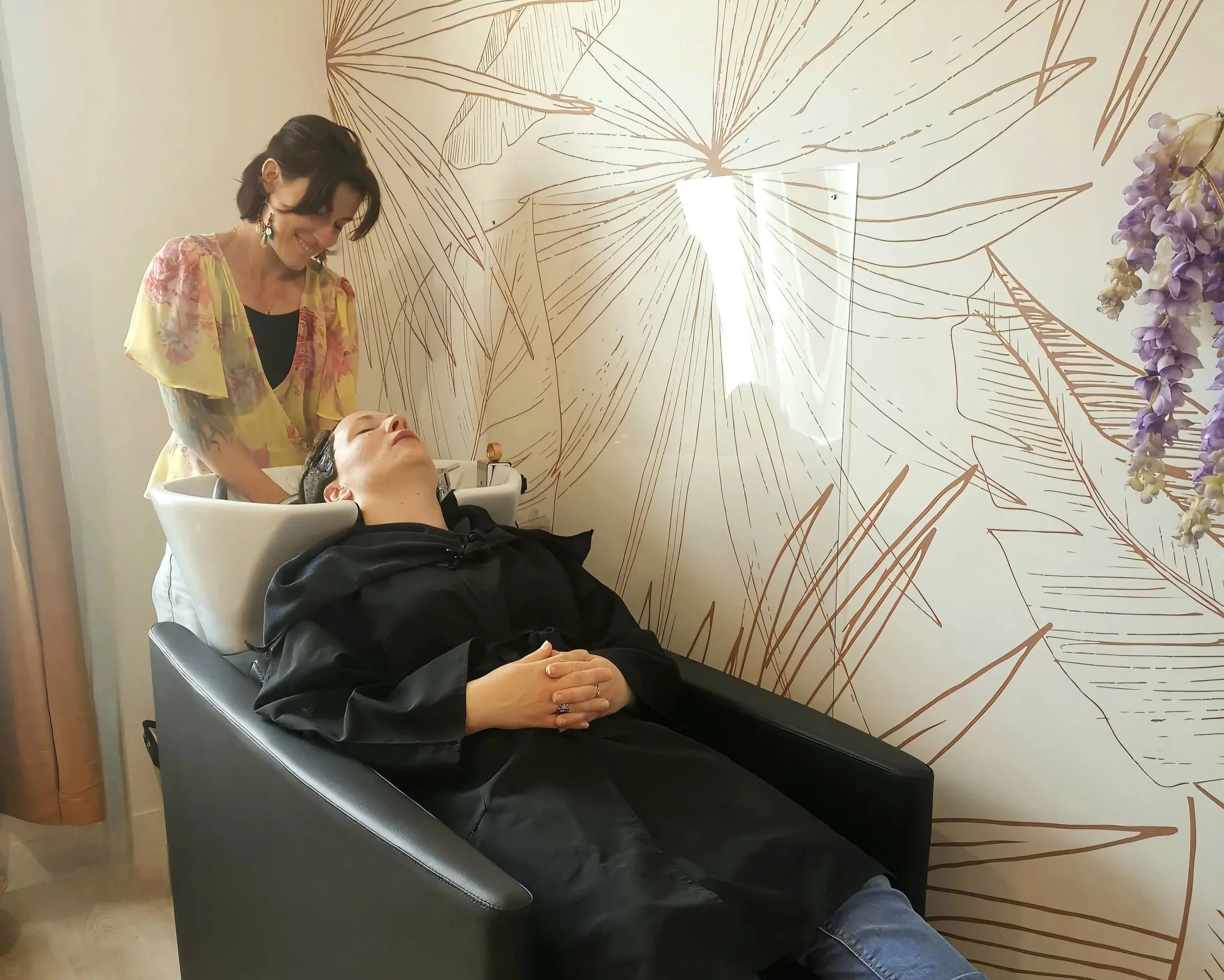 Une femme est allongée dans un fauteuil de spa ou centre de soins capillaires, une autre femme se trouve à côté, souriante, en train de lui laver ou de lui faire un soin capillaire. Le mur en arrière présente un design artistique avec des lignes représentant des feuilles ou des fleurs, et il y a des fleurs violettes en décoration sur le côté droit de l'image.