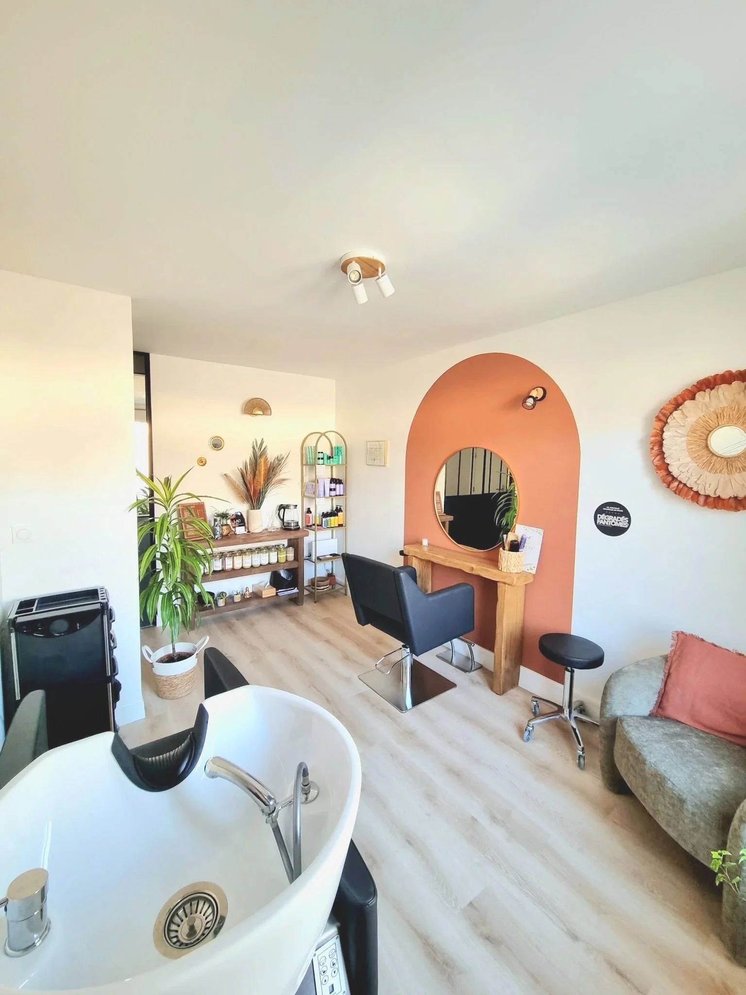 Salon avec un miroir rond, un fauteuil marron, un canapé gris, des plantes vertes, étagère avec des produits de beauté, décoration murale