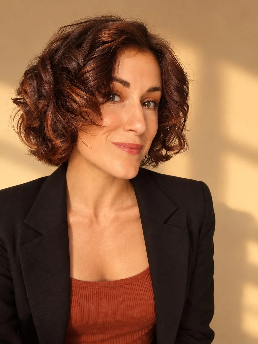 Une femme souriante avec des cheveux bouclés de couleur brun rouge, portant une veste noire et un haut orange, posant devant un fond beige
