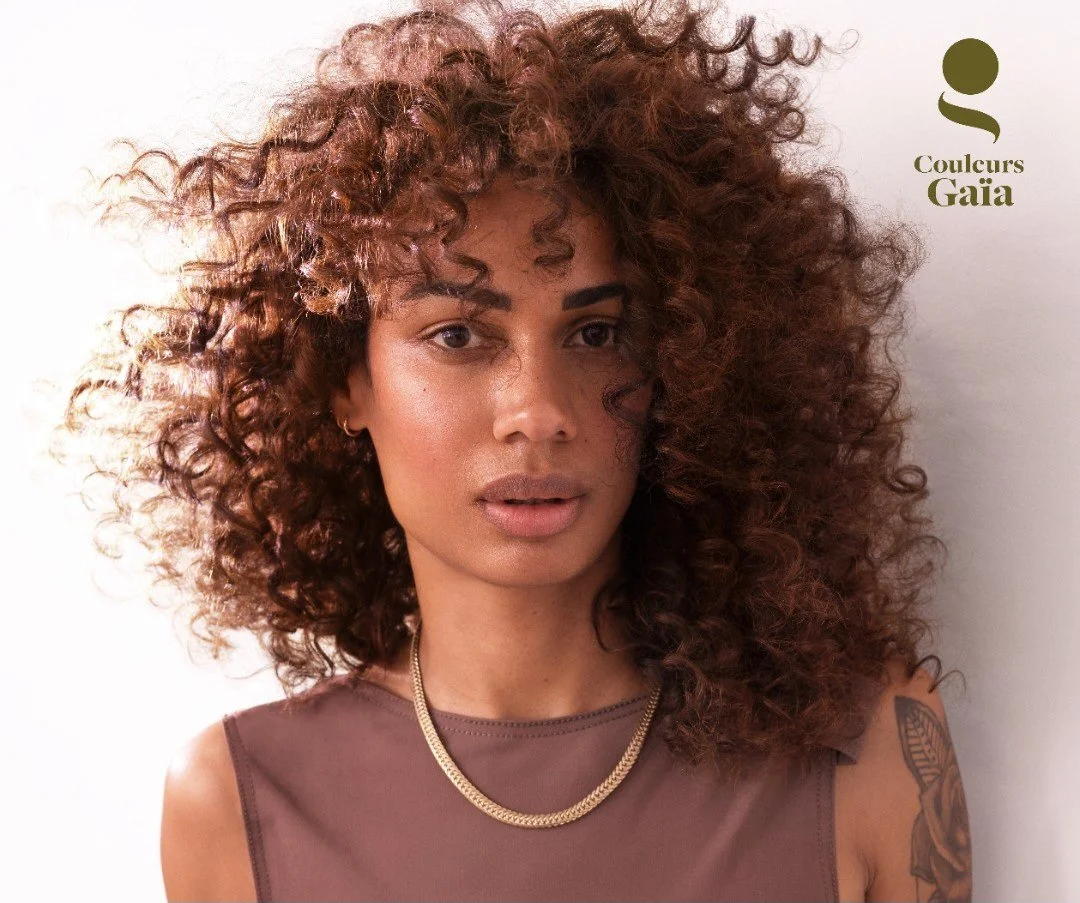 Portrait d'une femme avec des cheveux bouclés, portant un haut marron, une chaîne dorée et un tatouage sur l'épaule gauche, fond blanc, logo 'Couleurs Gaia' en haut à droite.