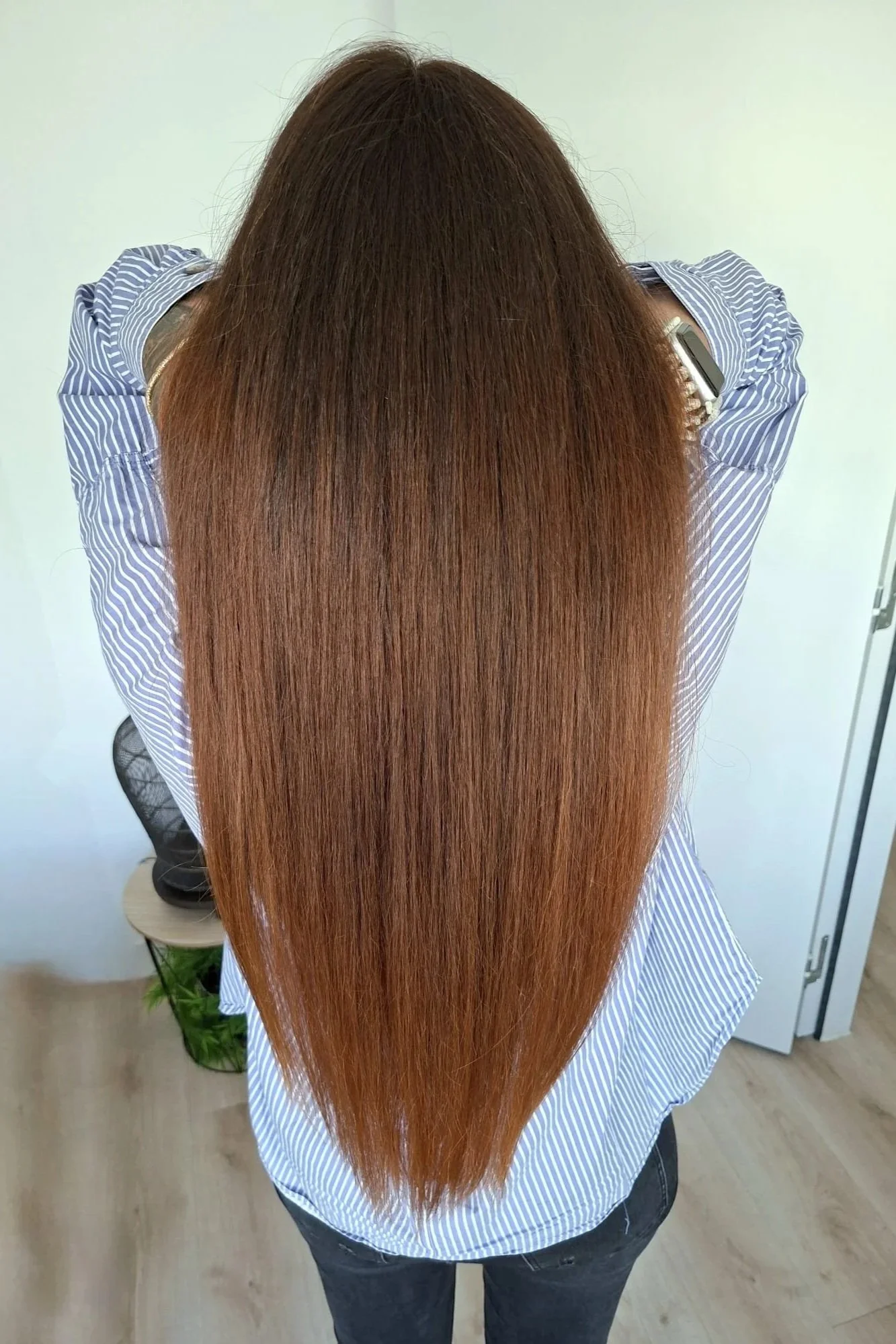 Une personne vue de derrière avec de longs cheveux roux brillants, portant une chemise à rayures bleues et blanches et un jean noir, en train de pencher la tête en avant.