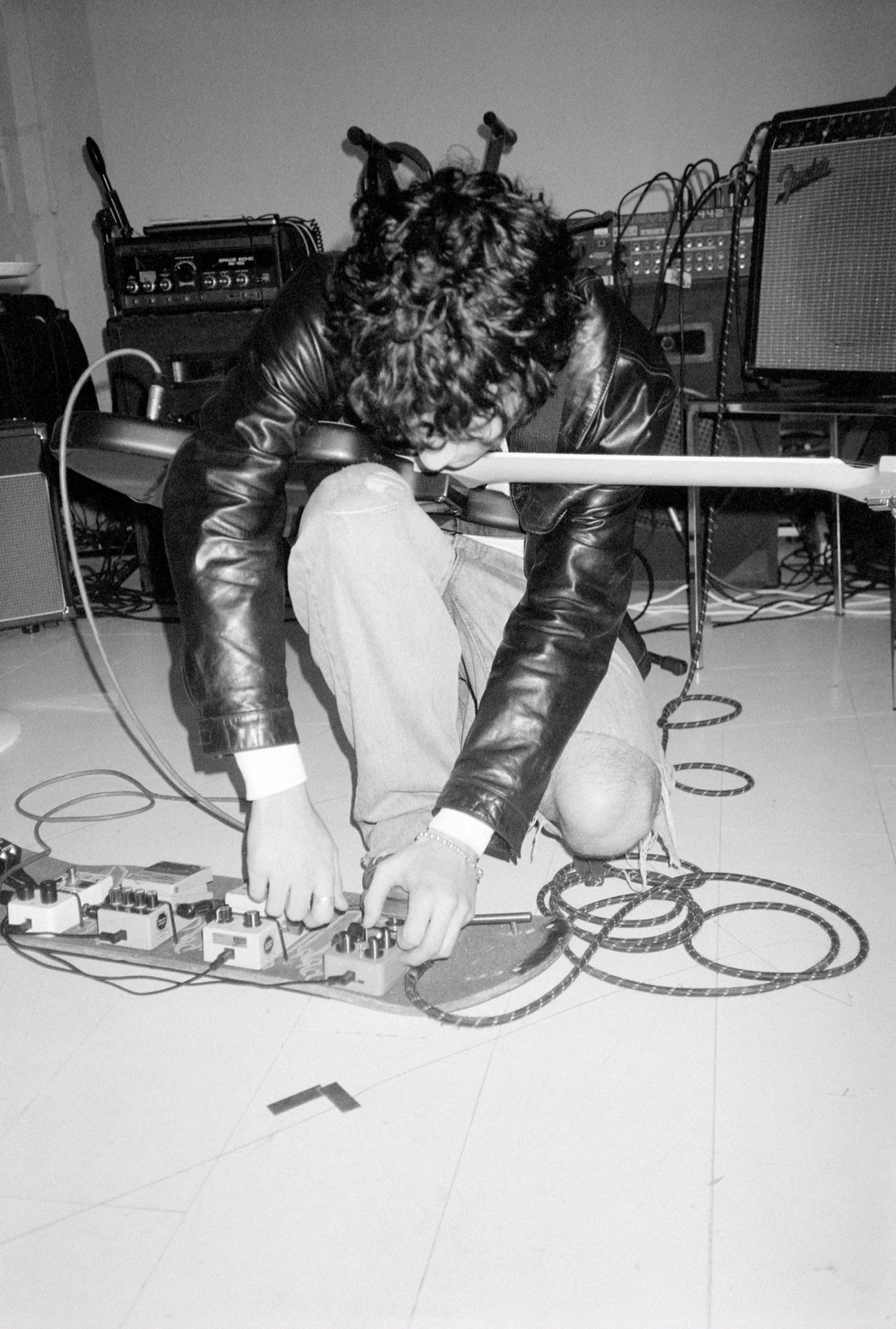 A person with curly hair wearing a leather jacket kneels on the ground, adjusting audio equipment connected with multiple cables in a room with musical and audio gear in the background.
