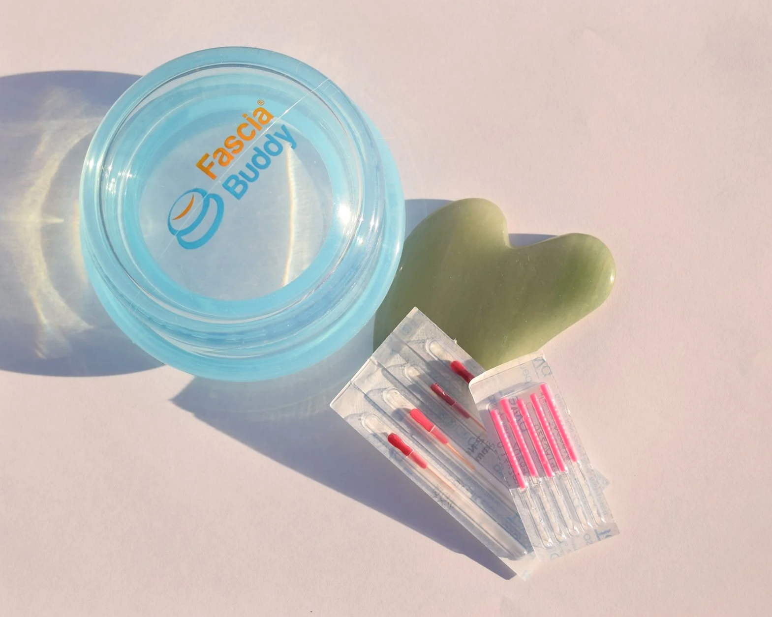 A green jade Gua Sha, a small plastic myofascial cup with a logo, and acupuncture needles with pink needles on a white surface.