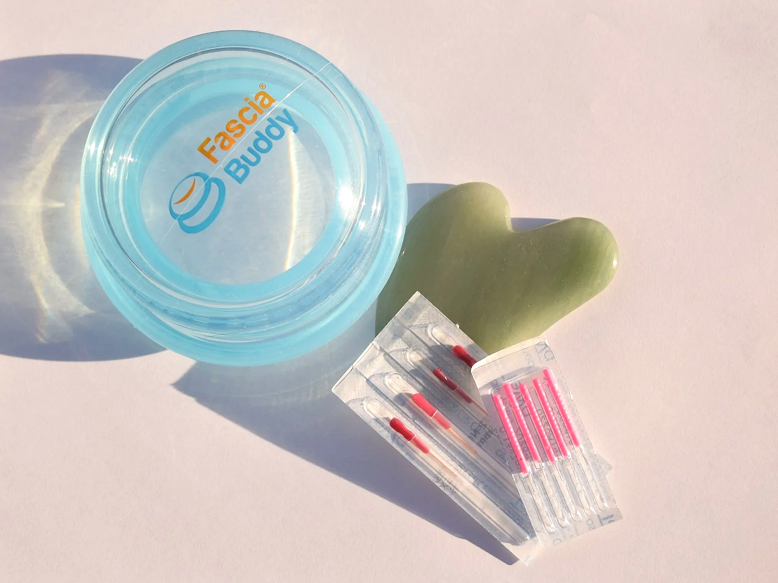A clear plastic container with the logo 'Fascia Buddy.' A green heart-shaped stone and a pack of acupuncture needles are placed on a white surface, casting shadows.
