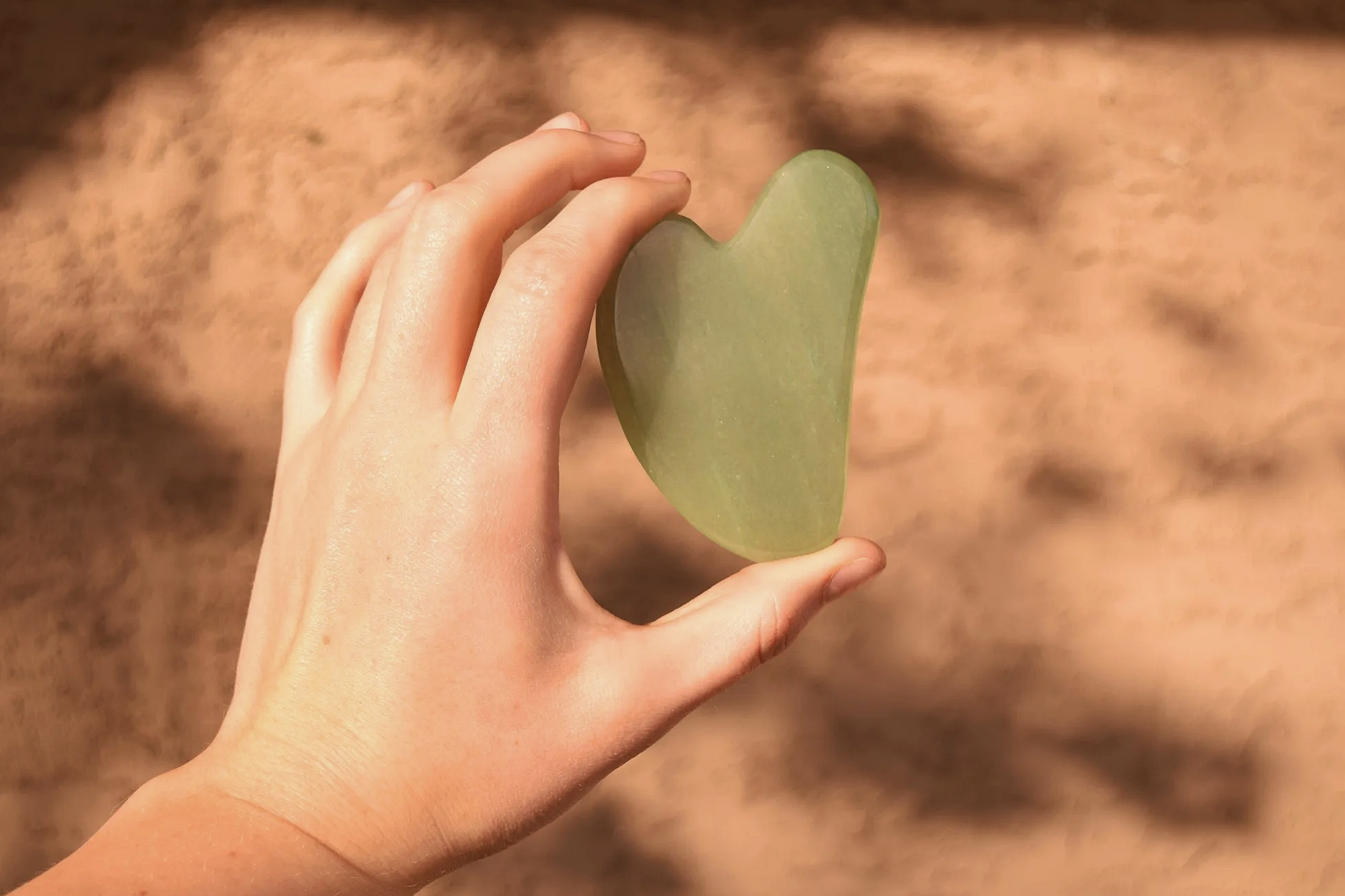 A hand holding a heart-shaped green jade Gua Sha against a blurred brown background.