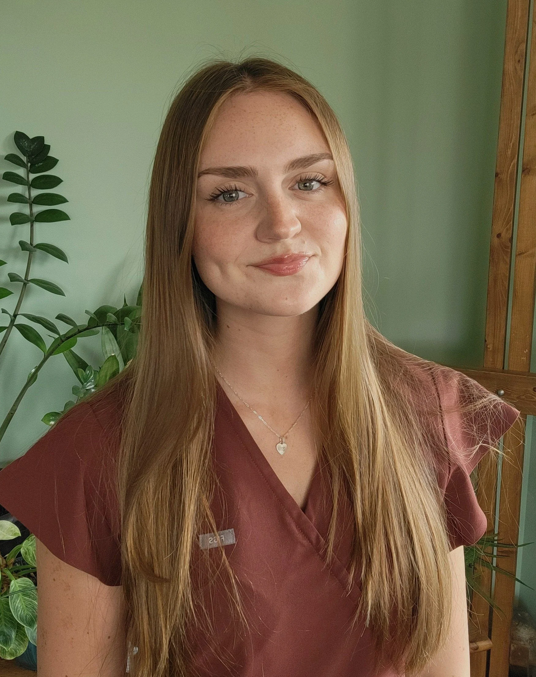A young woman with long, straight, light brown hair, wearing a maroon uniform top, standing in front of a green wall and a plant.