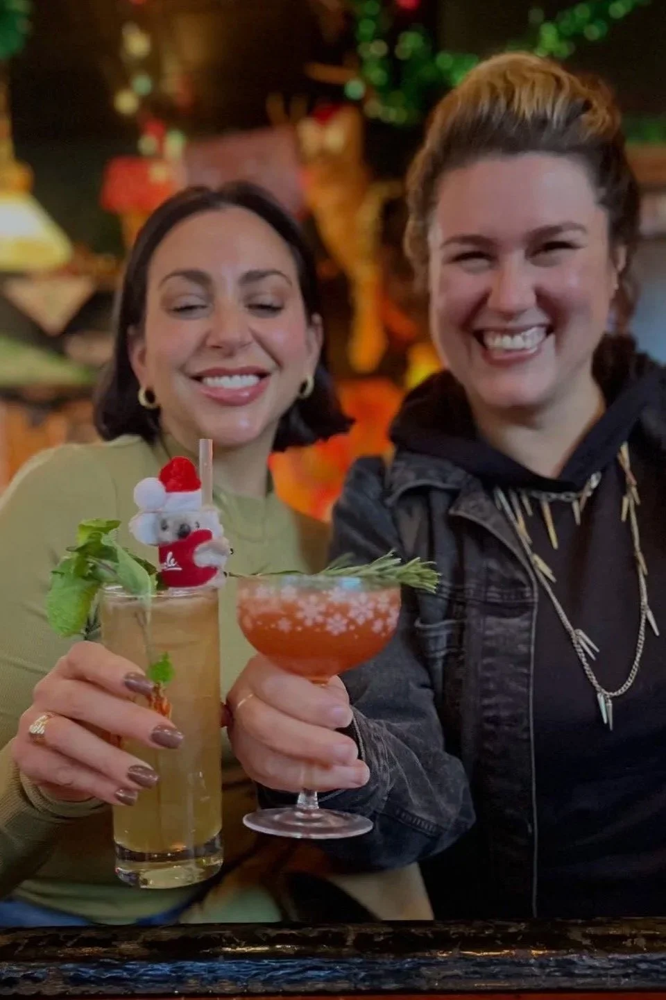 Two women smiling at a bar while holding colorful garnished cocktails.