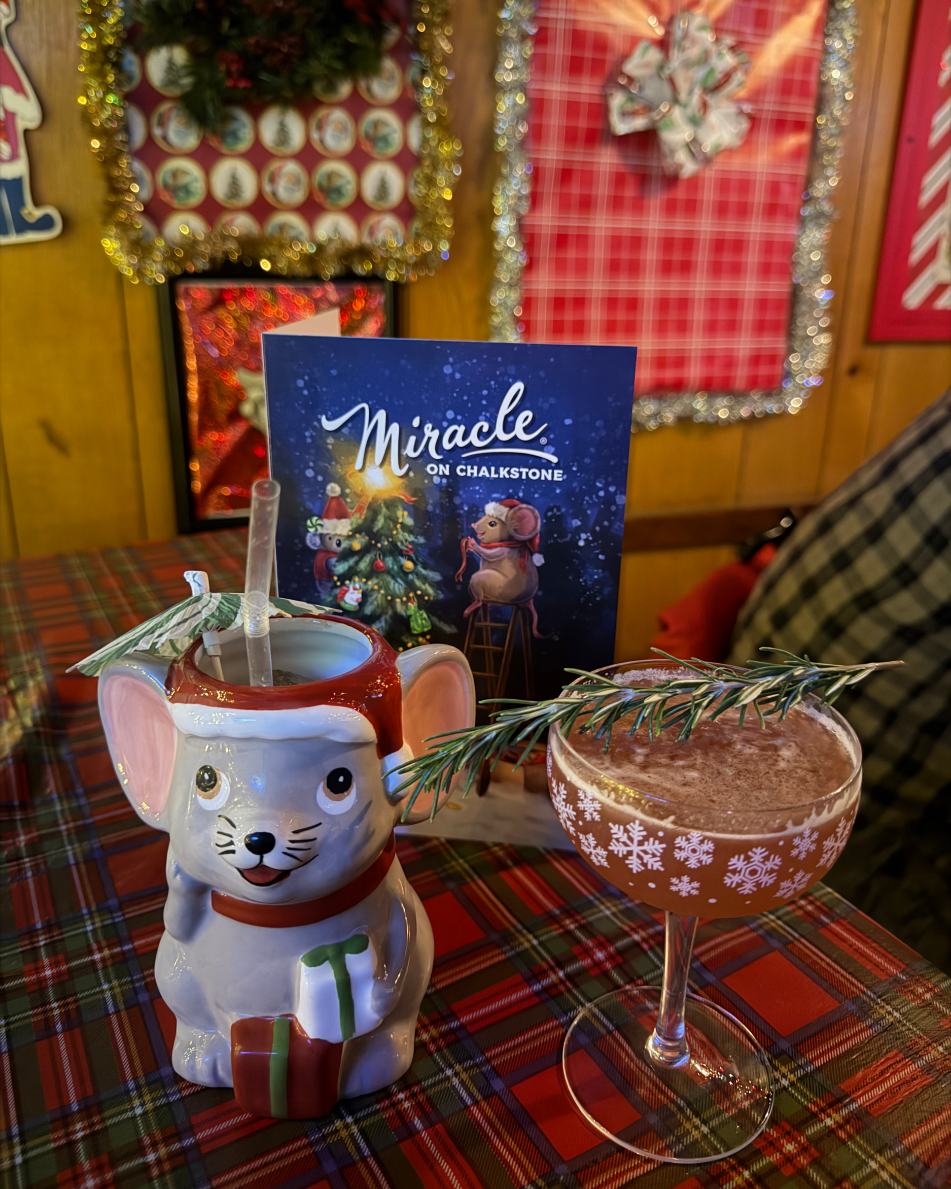 Festive Christmas table with a mouse-shaped Santa mug, snowflake cocktail garnished with rosemary, plaid tablecloth, and holiday decorations.