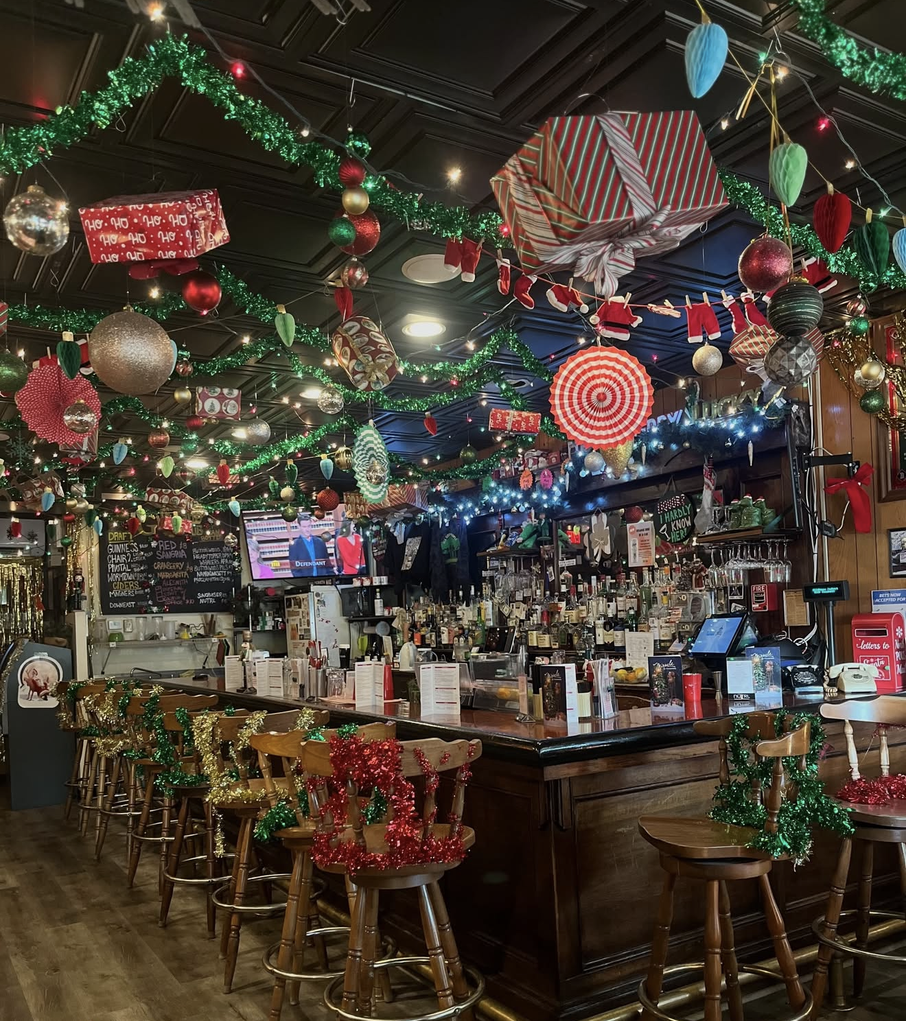 Festively decorated bar with Christmas ornaments, garland, string lights, a fully stocked liquor counter, and wooden bar stools.