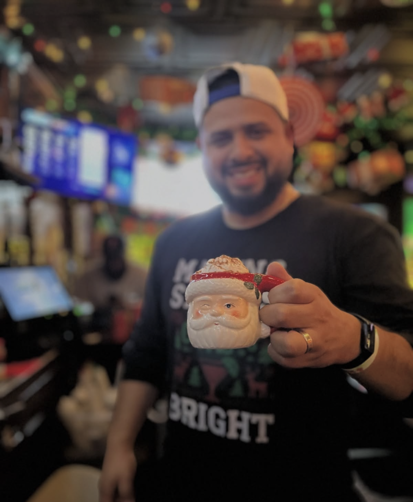 Smiling man in a backward baseball cap holding a Santa Claus mug in a festive bar or arcade.