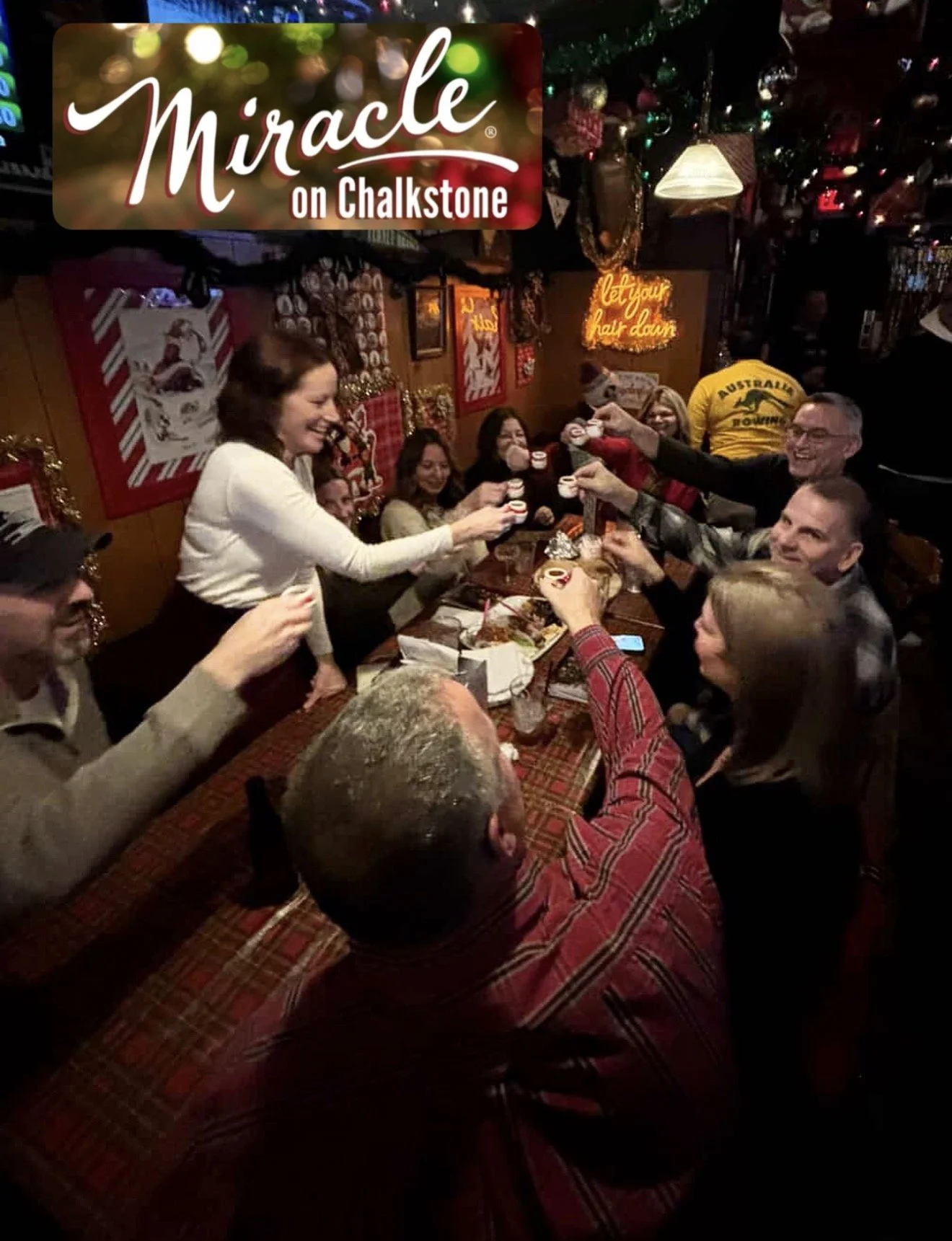 Group of people celebrating with small cups around a table inside a festive pub decorated for Christmas, with a “Miracle on Chalkstone” sign.