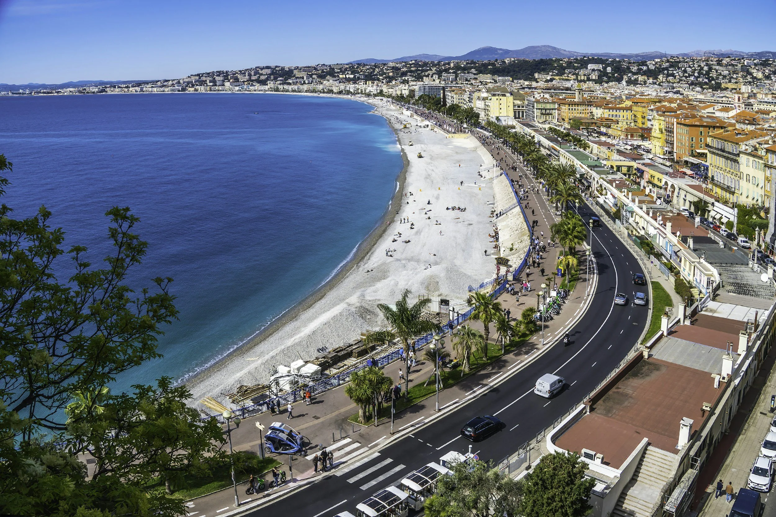 Spiaggia e promenade affiancata da un lungomare affollato di persone e palme, con edifici colorati e una strada trafficata.