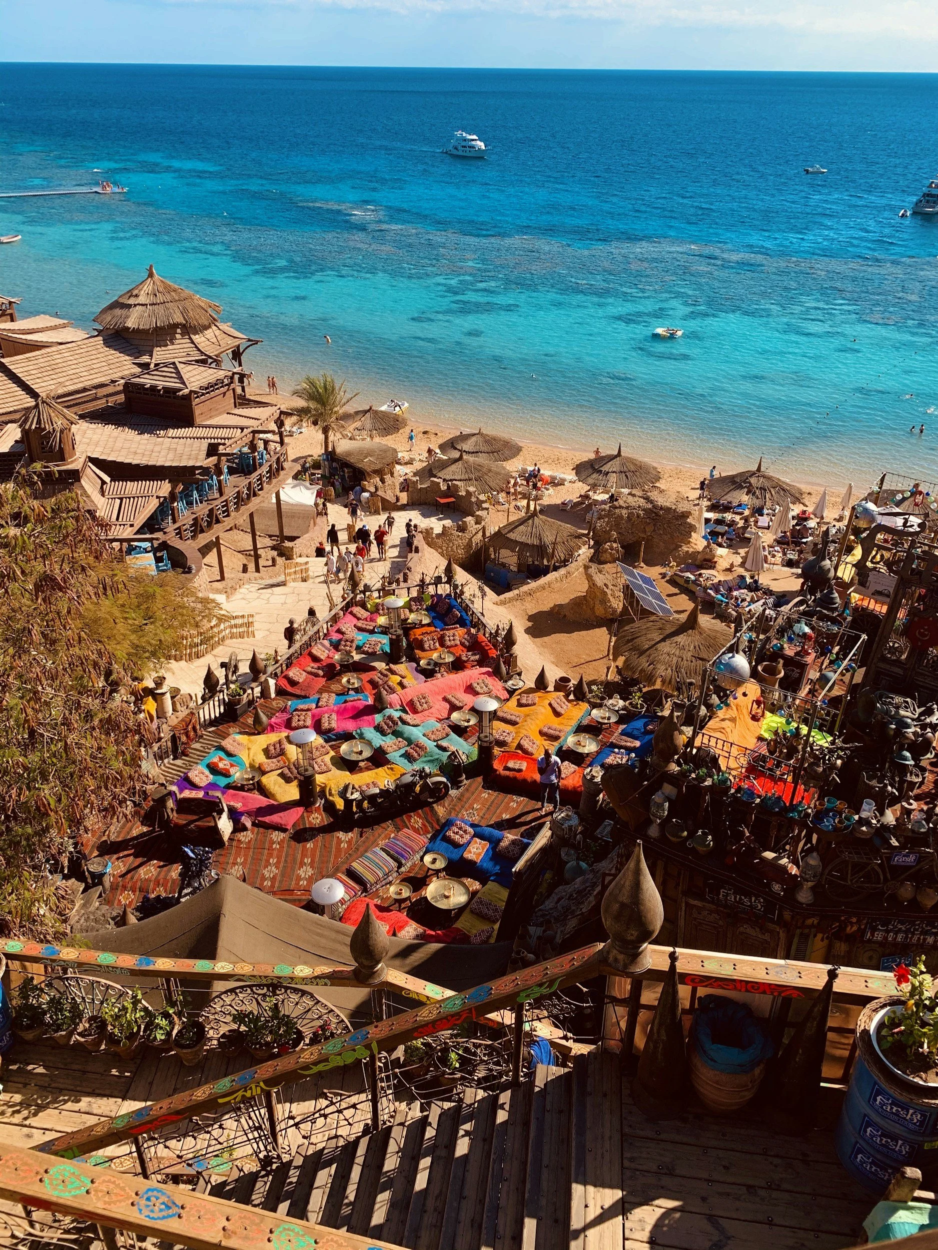 Vista di una spiaggia affollata con ombrelloni di paglia, persone che si godono il sole e l'acqua, barche in mare e un ristorante sulla spiaggia con tetto di paglia, tutto con vista sul mare azzurro e cielo sereno.
