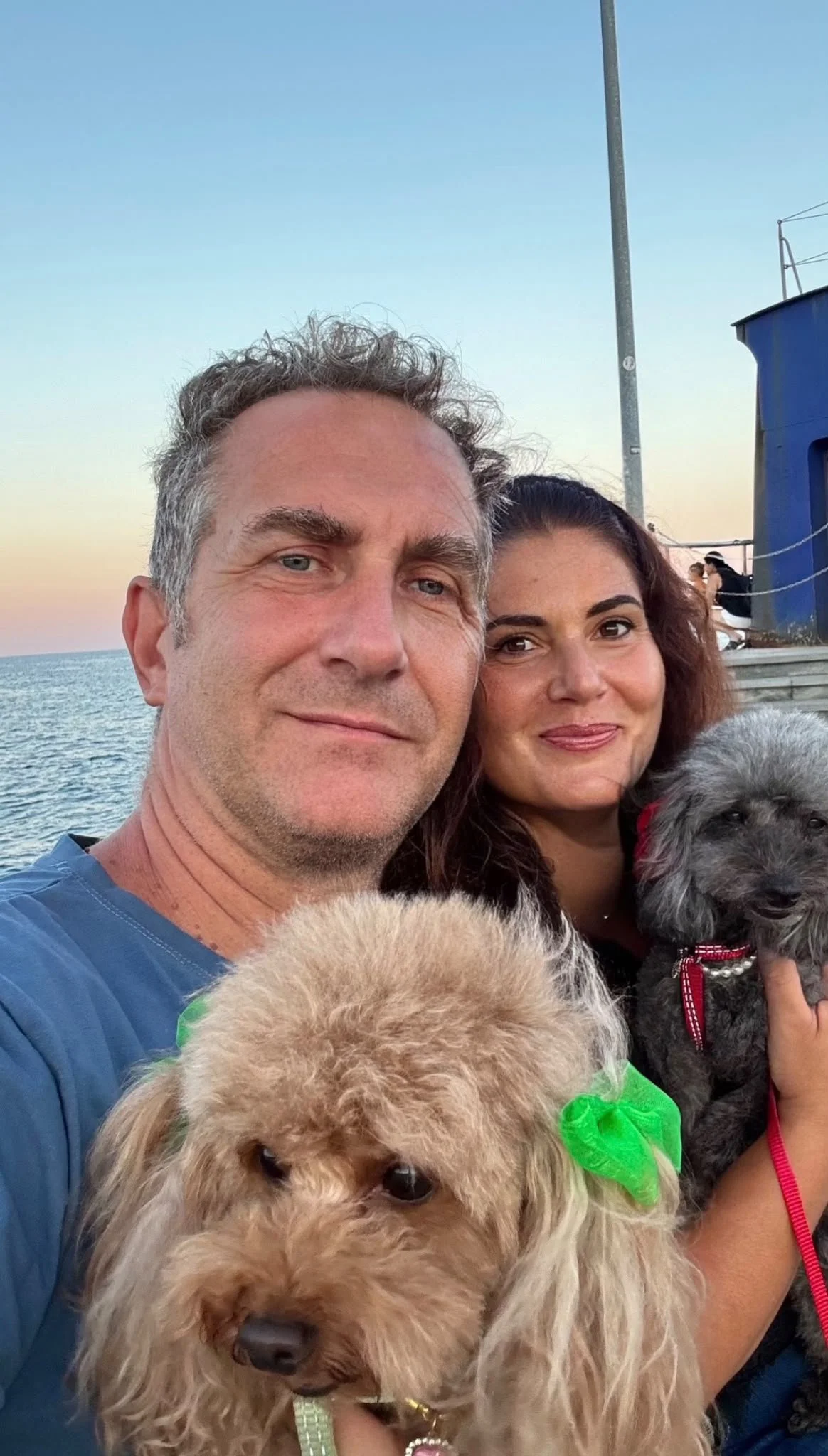 Una coppia sorride con due cani sotto il cielo al tramonto, vicino al mare.