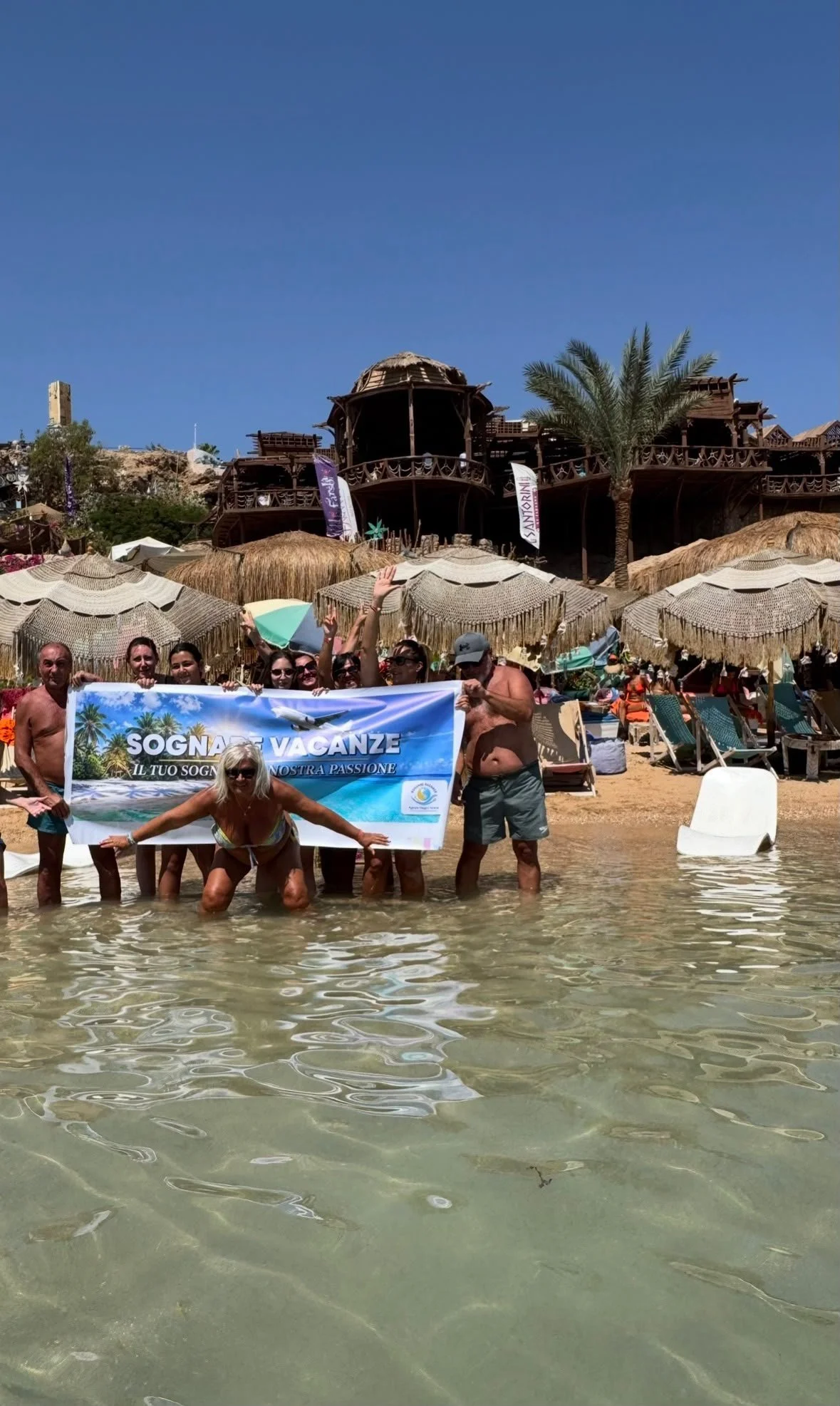 Gruppo di persone in spiaggia che tiene uno striscione con scritte in italiano, con sfondo di ombrelloni di paglia, palme e una struttura in legno, sotto un cielo azzurro.