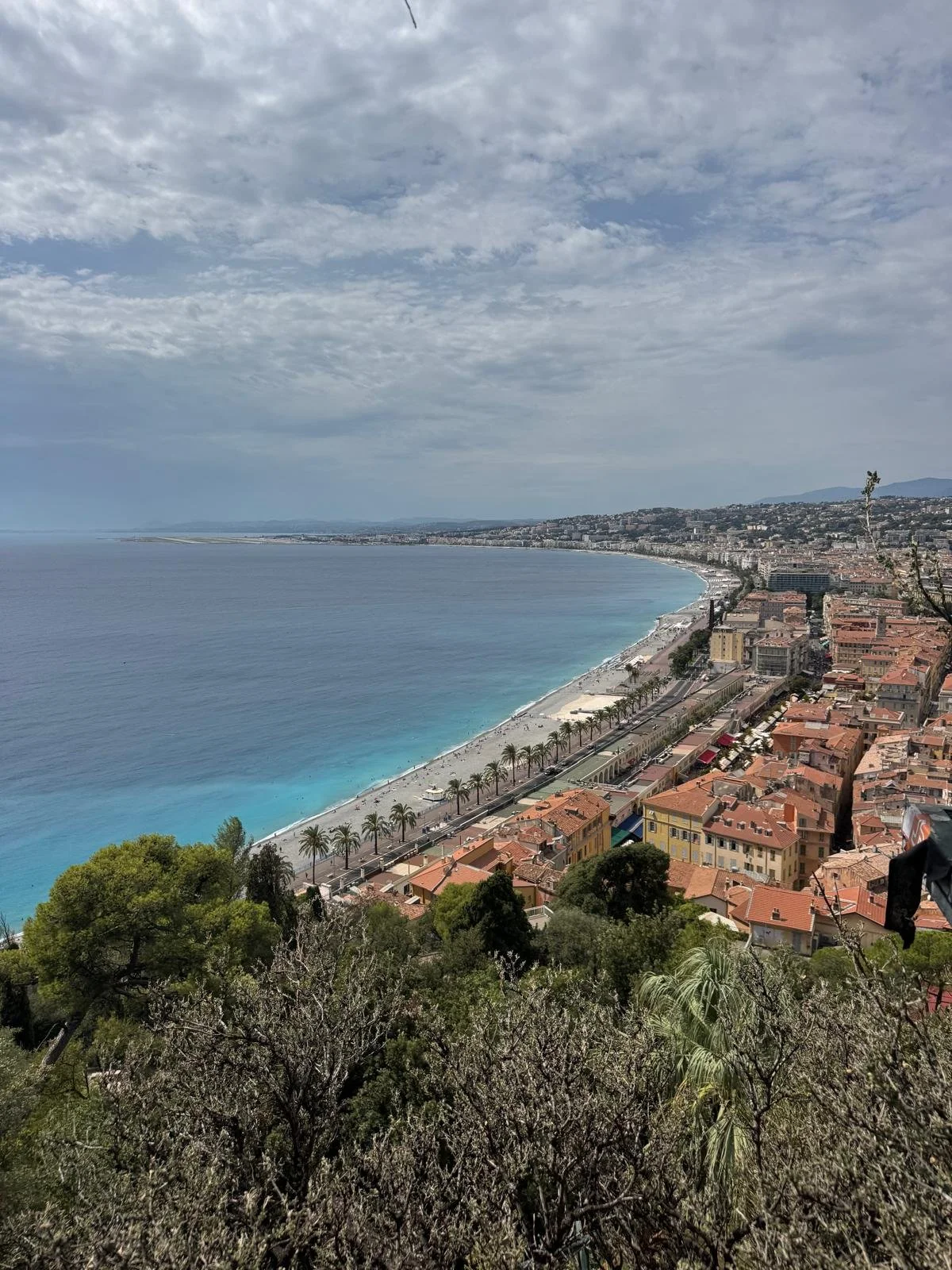 Vista panoramica di una spiaggia lungo la costa con alberi di palma, case e un mare azzurro sotto un cielo parzialmente nuvoloso