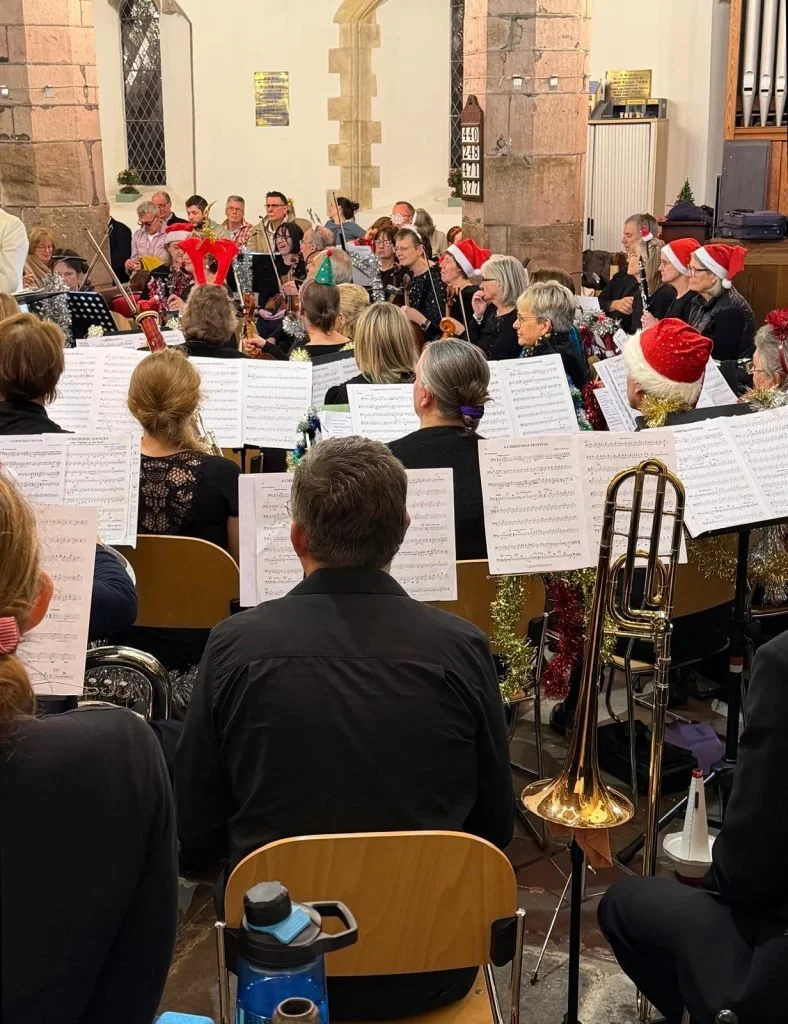A Christmas concert taking place in a church with musicians wearing holiday accessories like Santa hats and reindeer antlers. Some musicians are playing string and wind instruments, with sheet music in front of them.