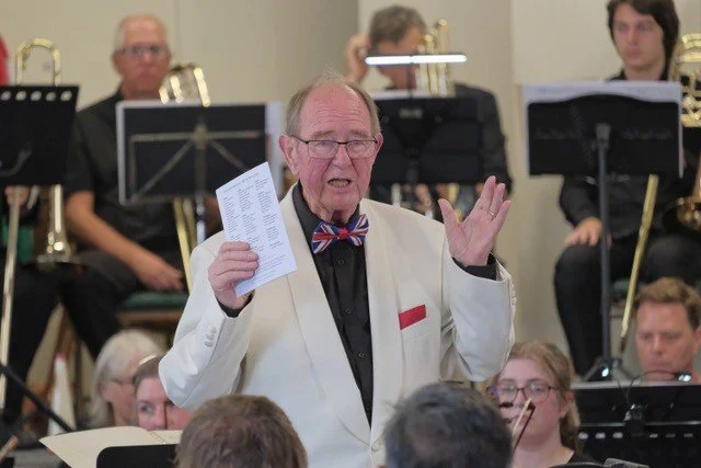 An elderly man in a white tuxedo with a bow tie, conducting an orchestra during a performance.