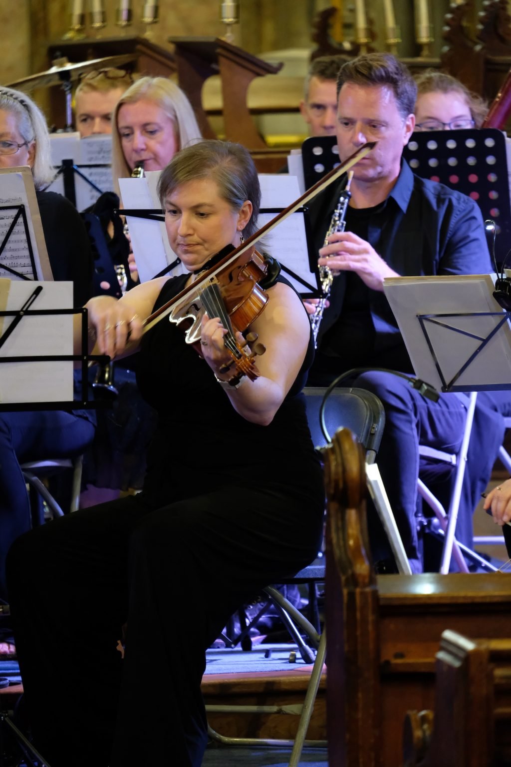 An orchestra performing inside a church or concert hall, with a woman playing the violin in the foreground and other musicians behind her.