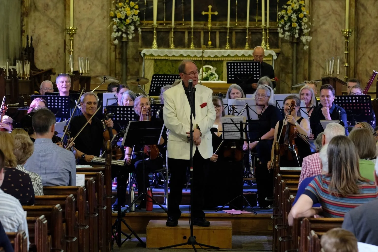 Orchestra seated behind a man in a white blazer speaking into a microphone in a church or cathedral setting with an audience watching.