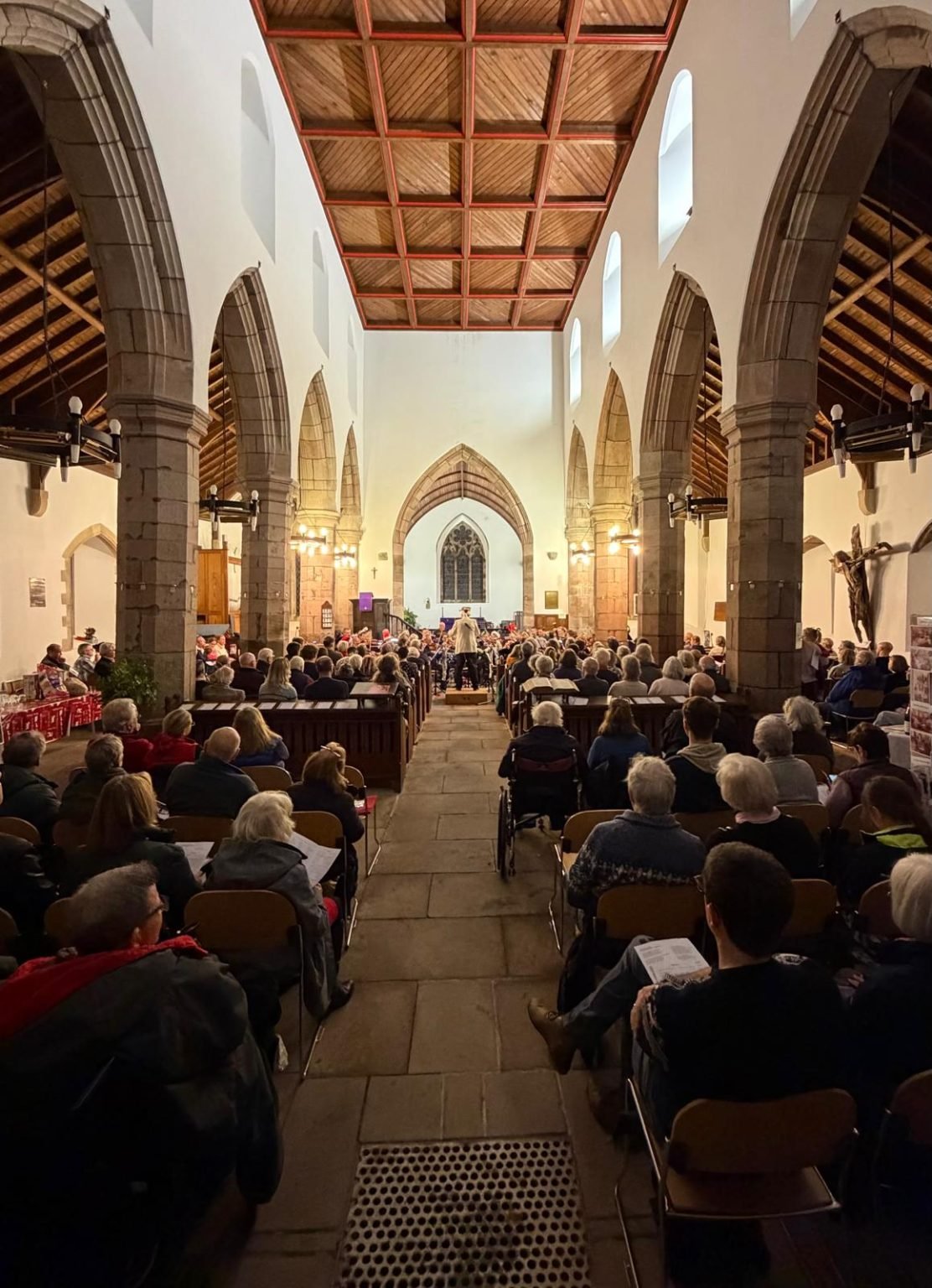 Inside a church filled with people attending a service or event, with rows of benches, stone arches, a high wooden ceiling, and a stained glass window at the front.