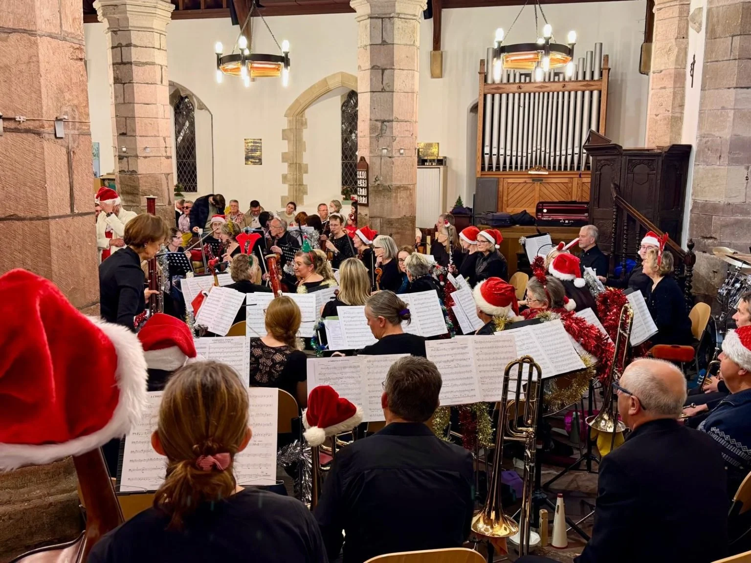 A large group of people performing Christmas music in a church, with some wearing Santa hats and festive decorations, during a holiday concert.