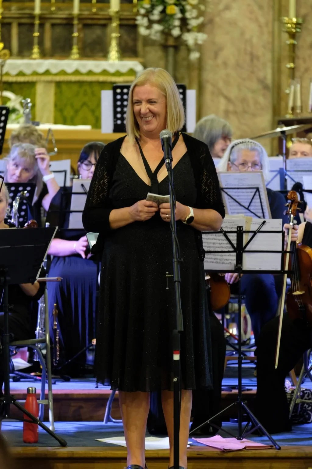 A woman with blonde hair in a black dress smiling and speaking into a microphone during a concert, with an orchestra playing behind her in a church or concert hall.