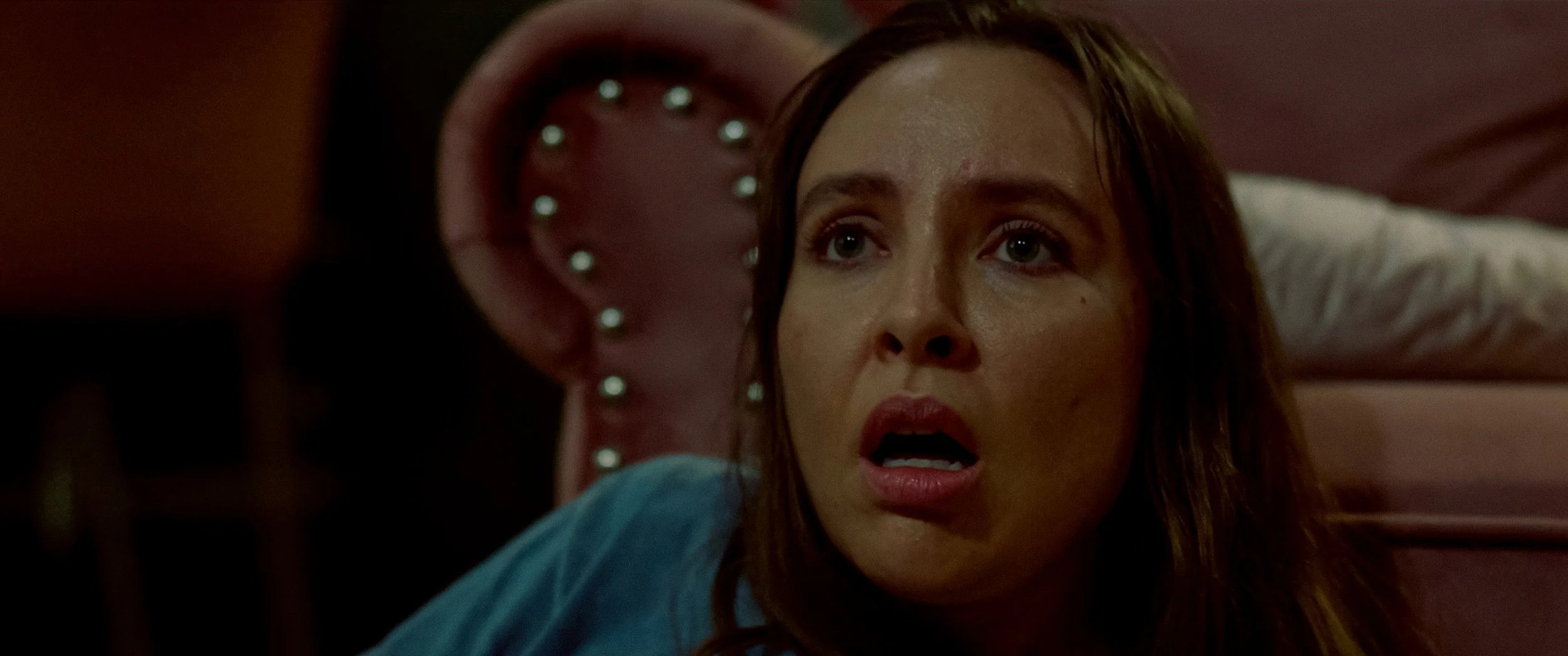 A woman with long brown hair lying in bed with a surprised or shocked expression, looking towards the right side of the image, with a pink tufted headboard and a pillow behind her.