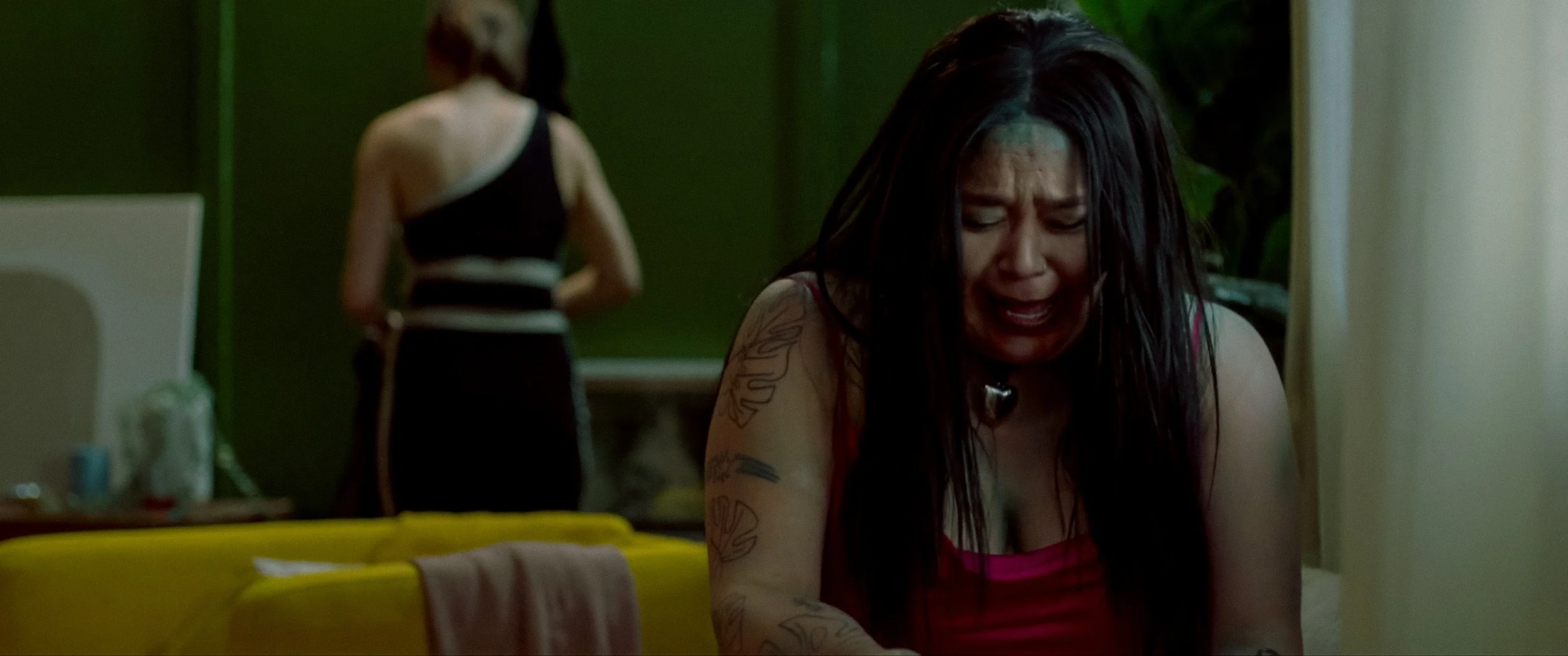 A woman with long dark hair, tattoos, and a distressed expression crying inside a room, with another woman standing in the background facing away.