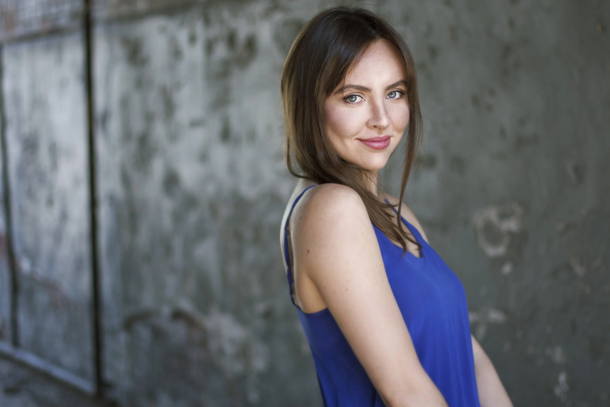 Bronwyn Szabo, a woman with fair skin and blue eyes wearing a blue sleeveless top, standing outdoors against a blurred concrete or stone wall.