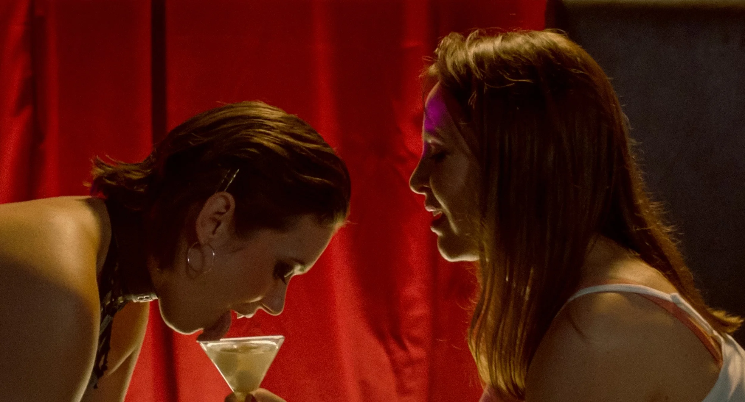Two women are leaning their foreheads together, with a red curtain as the background. One woman is holding a cocktail glass, and both women are smiling softly, engaged in an intimate moment.