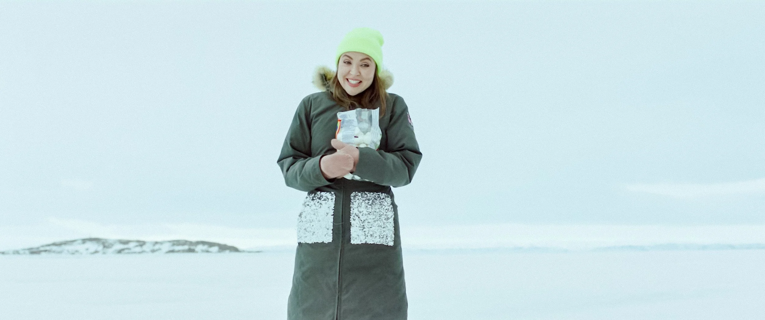 Woman standing in snowy landscape wearing a green beanie and a dark winter coat, holding a bag of marshmallows, smiling.