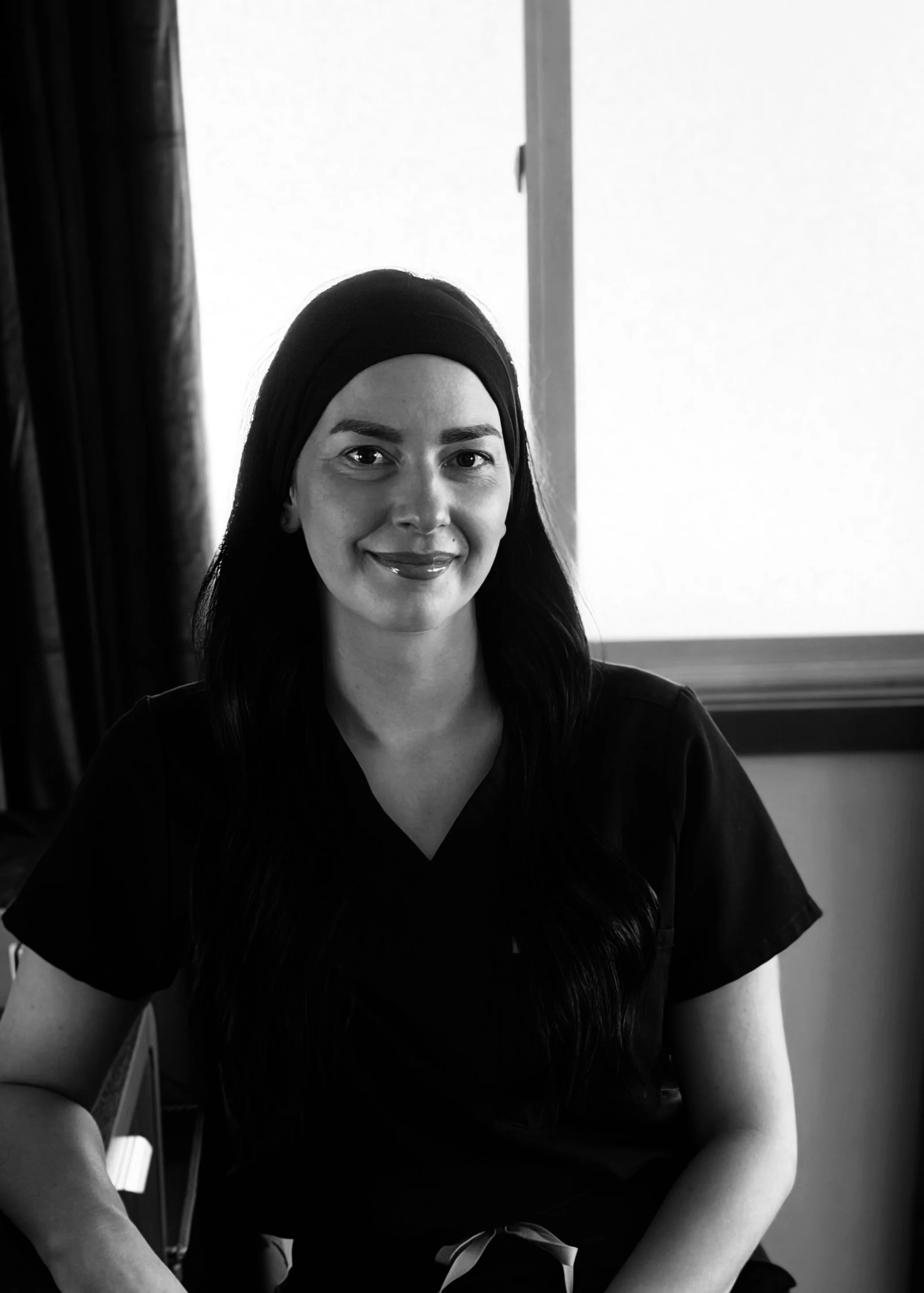 Smiling woman wearing a dark headband and black shirt, seated indoors by a bright window in a black-and-white portrait.