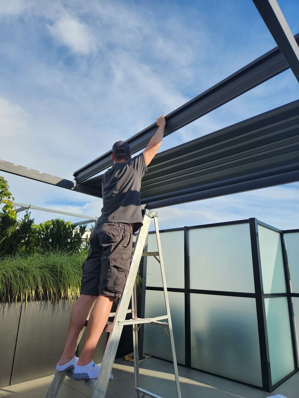Matt on a ladder reaching up to assemble a pergola frame on a rooftop patio