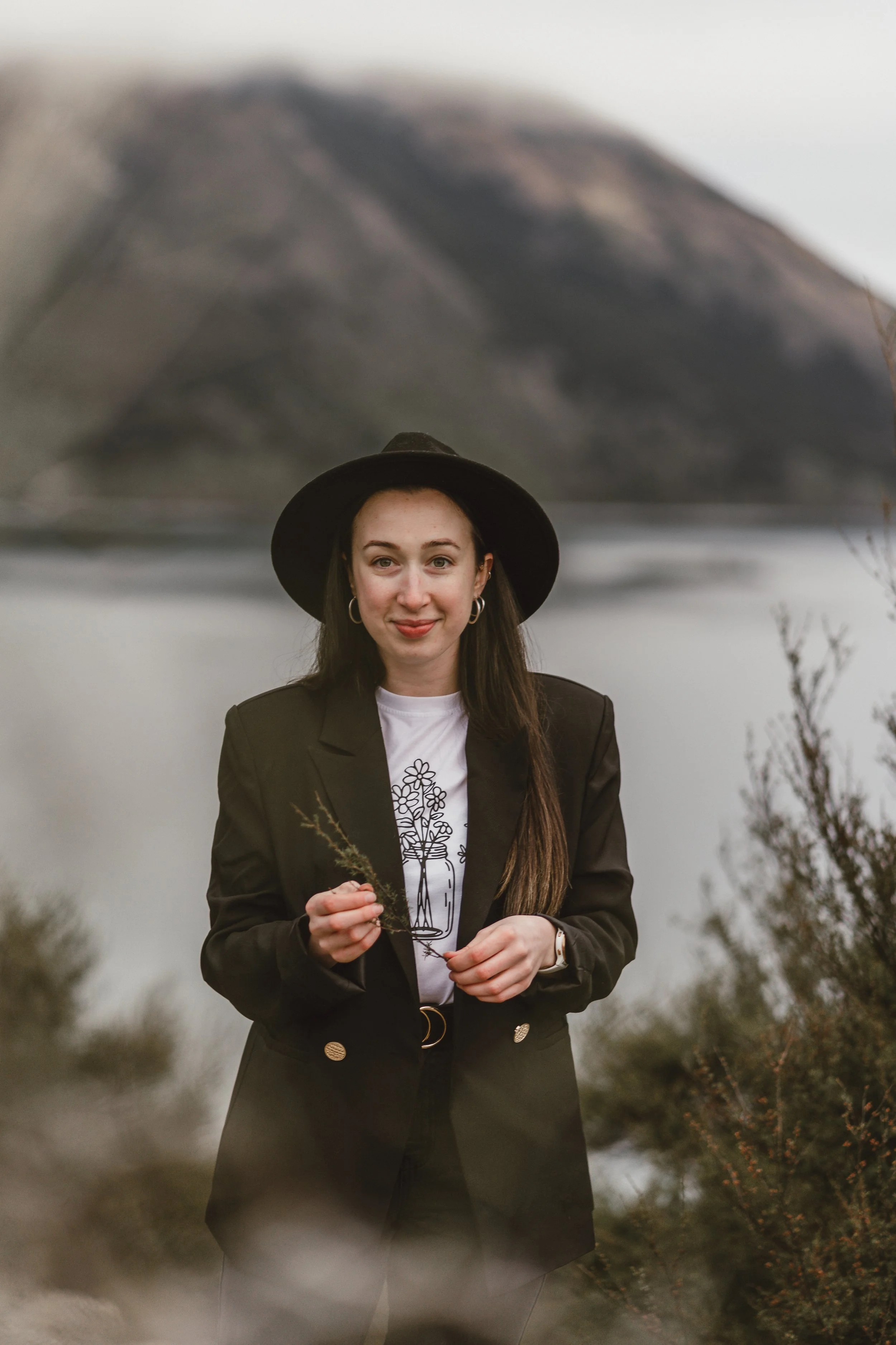 A young woman standing outdoors in a rural area with mountains in the background. She is wearing a black hat, earrings, a black blazer, and a white t-shirt, and is holding a small branch in her hand. The sky is overcast and the landscape is slightly blurred.