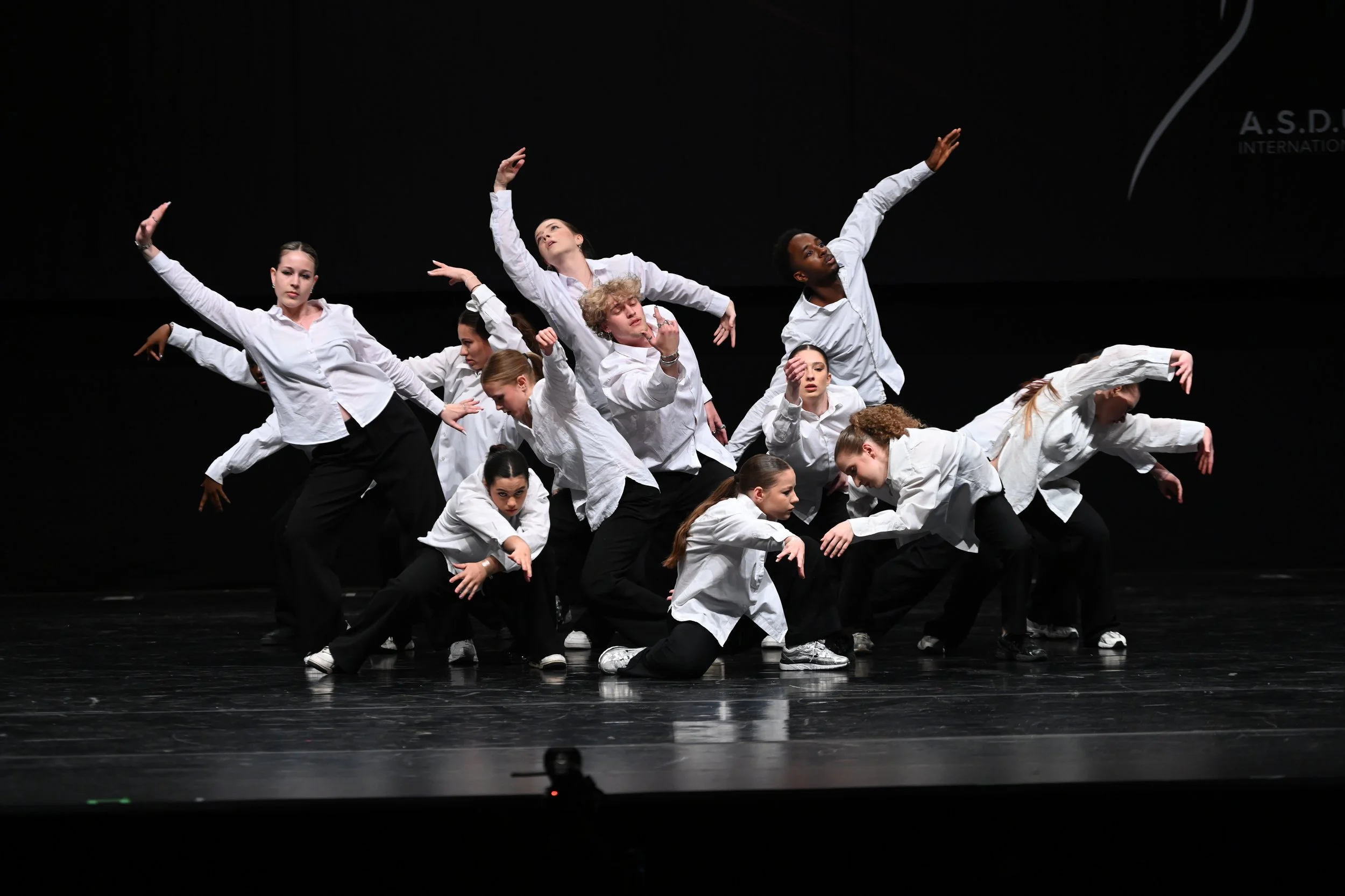 Tanzgruppe führt eine choreografierte Tanzroutine auf einer Bühne in weißen Hemden und schwarzen Hosen auf, während der Hintergrund dunkel ist.