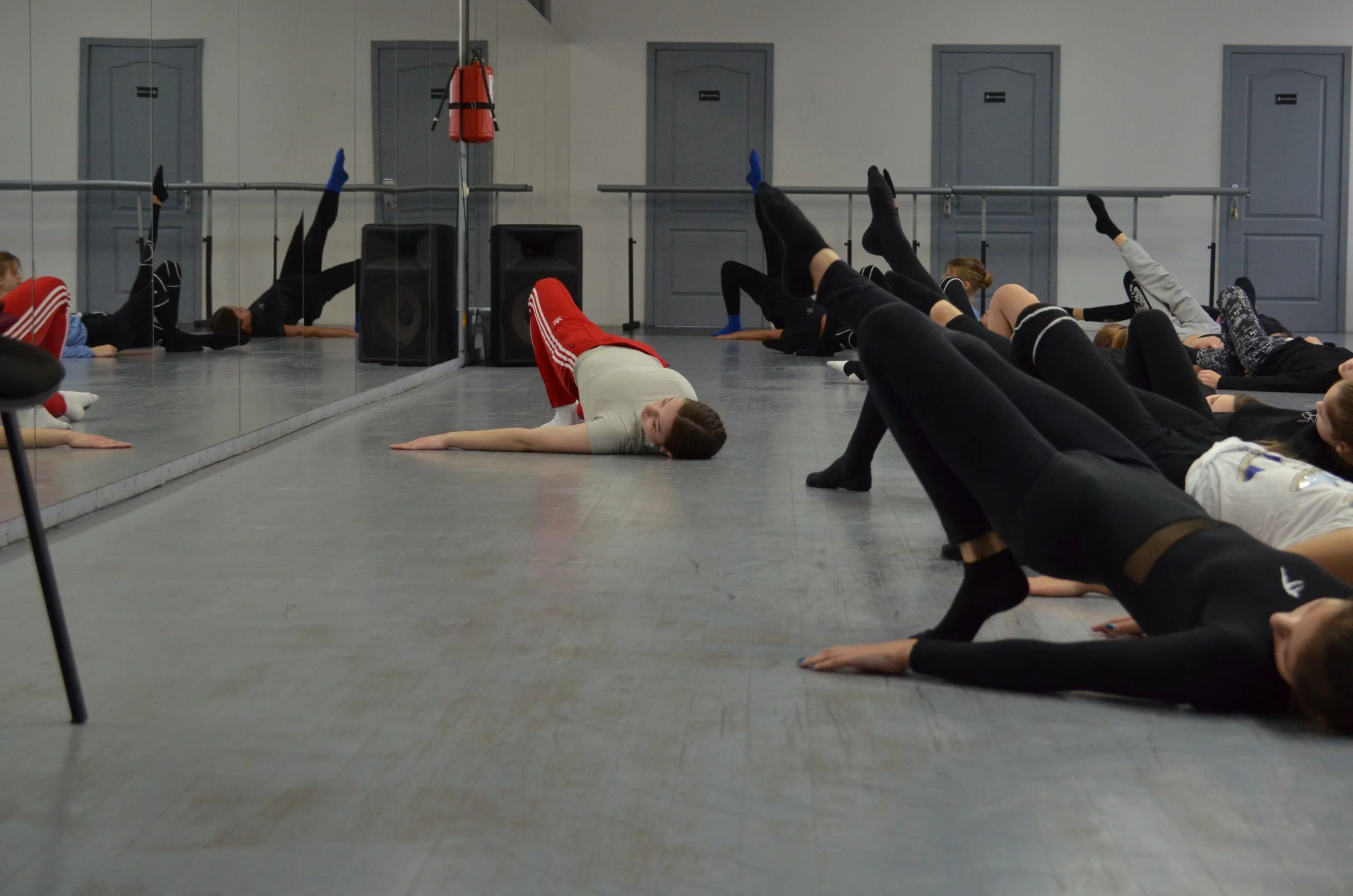 Gruppen von Menschen machen Dehnübungen auf dem Boden im Tanzstudio, die in eine Wand mit Spiegeln schauen.