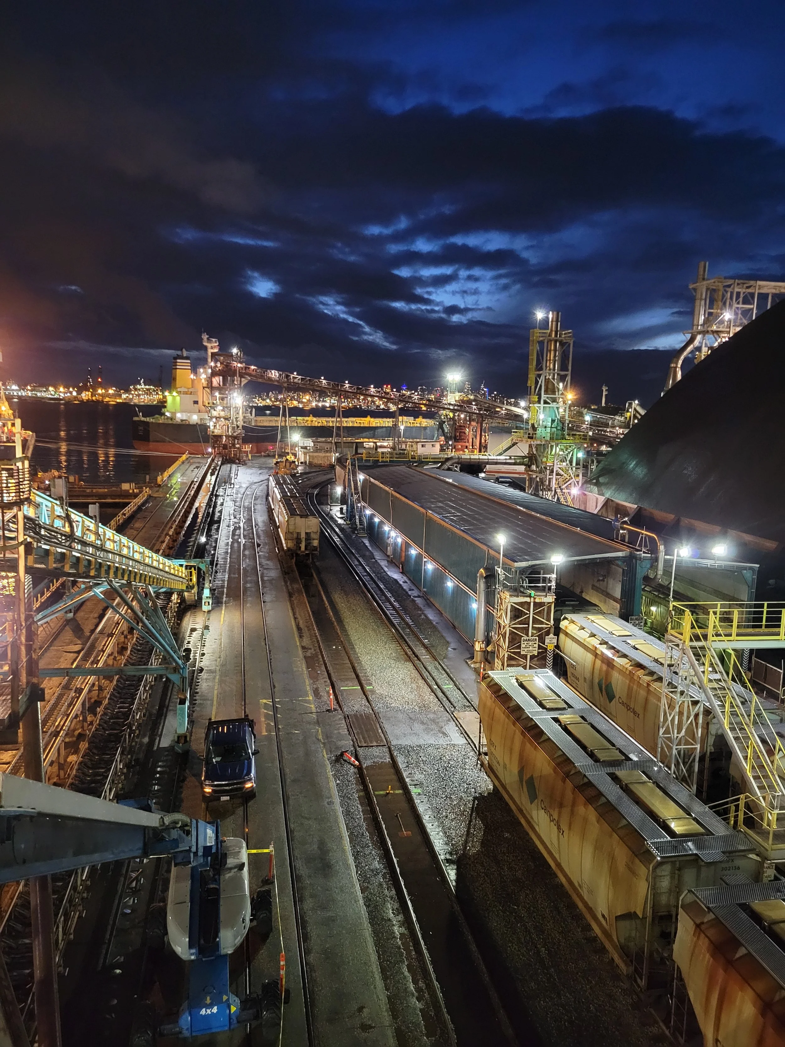 Nighttime scene at an industrial site with trains, tracks, and large machinery under a dark, cloudy sky with city lights in the background.