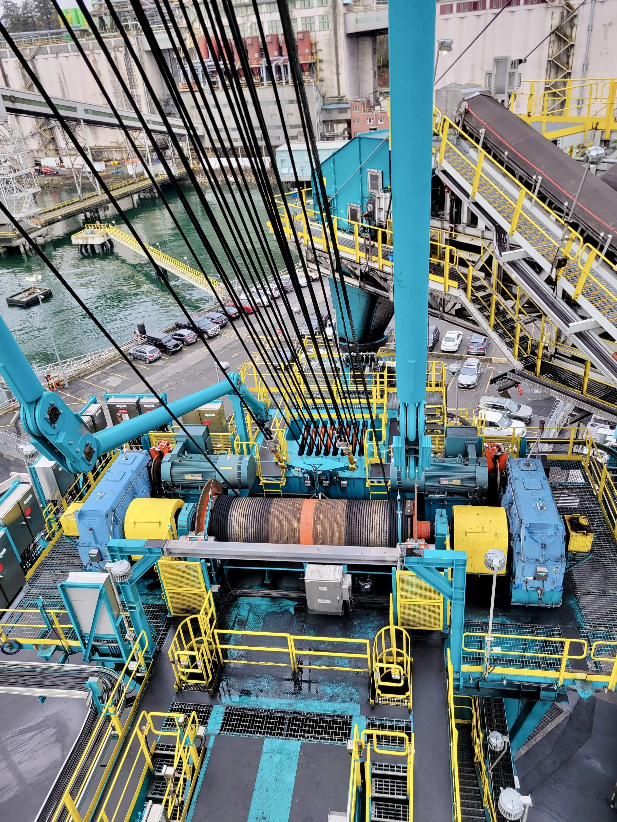 Industrial equipment with blue, yellow, and black components, including large rollers and a conveyor system, on a deck overlooking a parking lot and waterway.