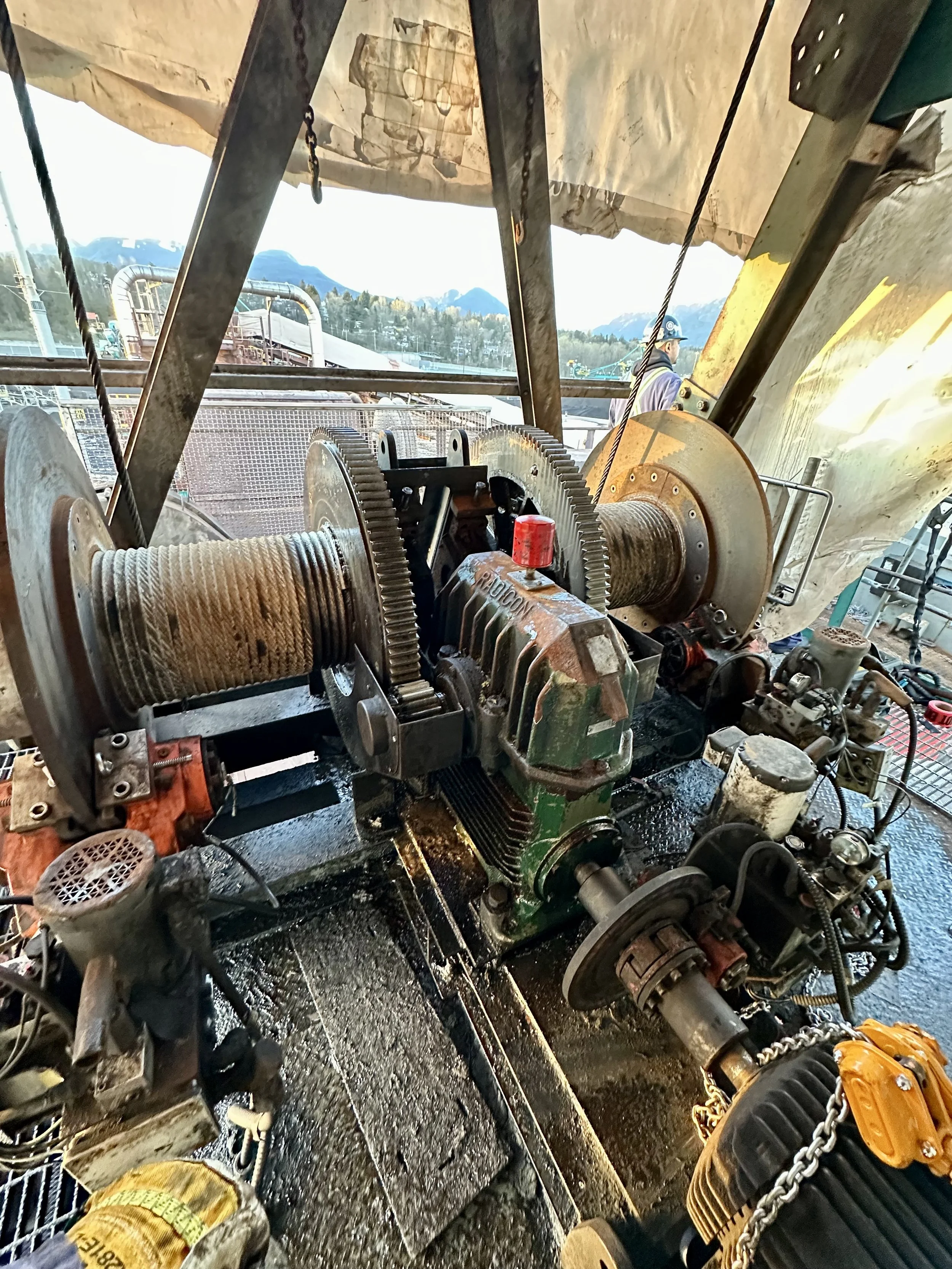 Construction worker in yellow gloves using a chainsaw to cut through metal in an industrial setting with machinery and mountains in the background.