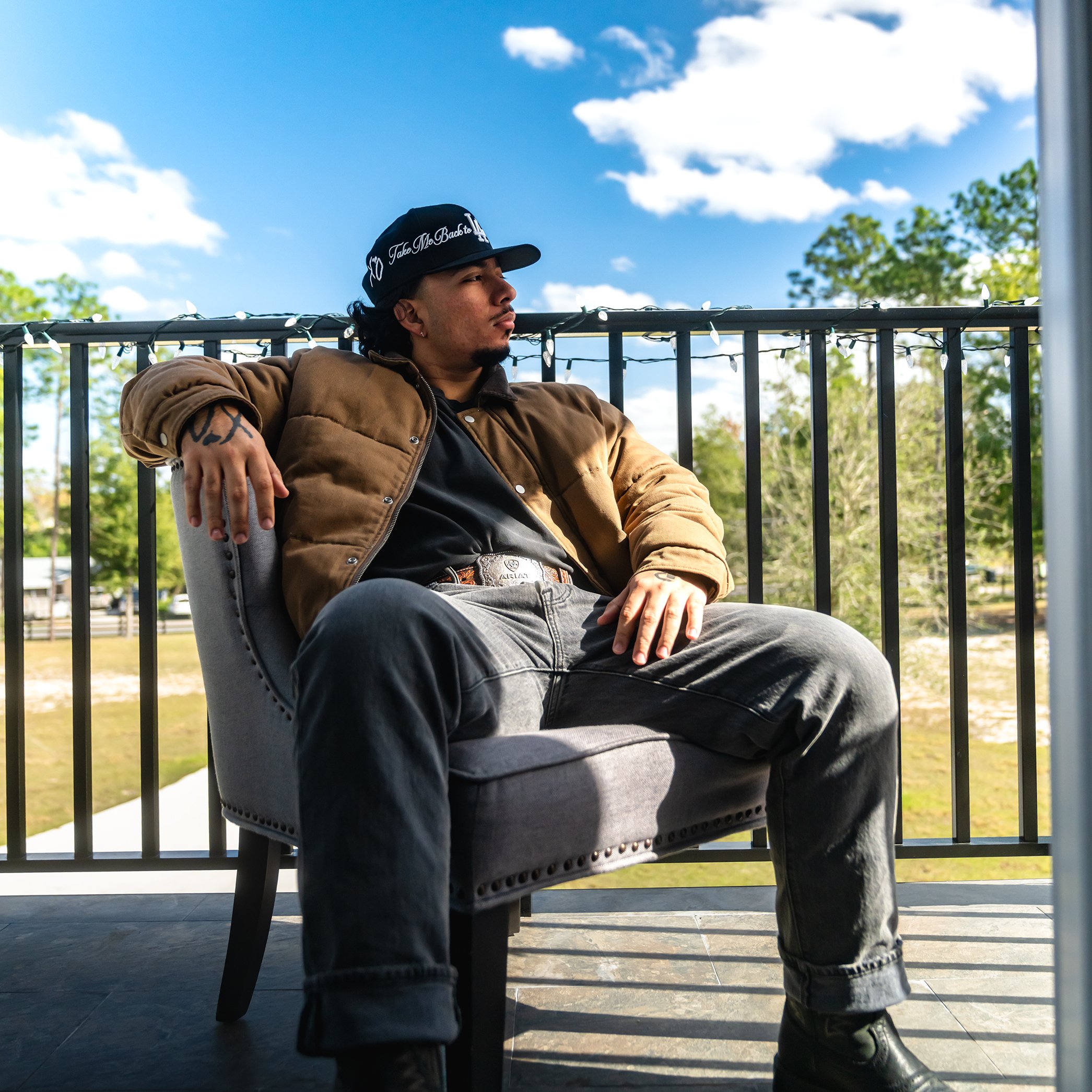 A young man with tattoos on his arm and a goatee, wearing a black cap with white writing, a brown jacket, black shirt, blue jeans, and boots, sitting on a bench outdoors on a sunny day with a clear blue sky and some clouds, trees, and a parking lot in the background.