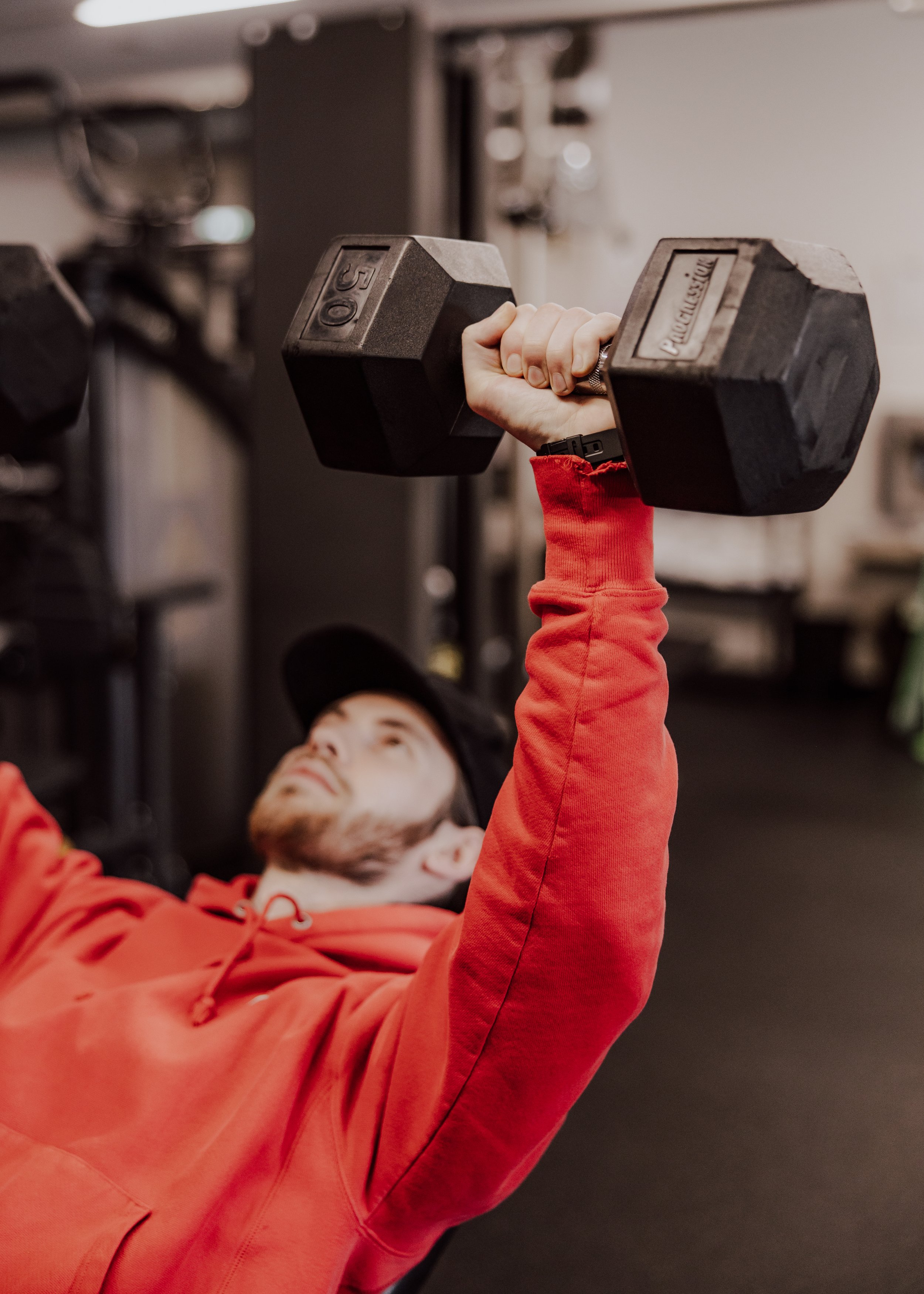 A person in a red hoodie is lying on the floor, holding a 50-pound dumbbell with their right hand raised above their head. The background appears to be a gym, with shelves and equipment visible.