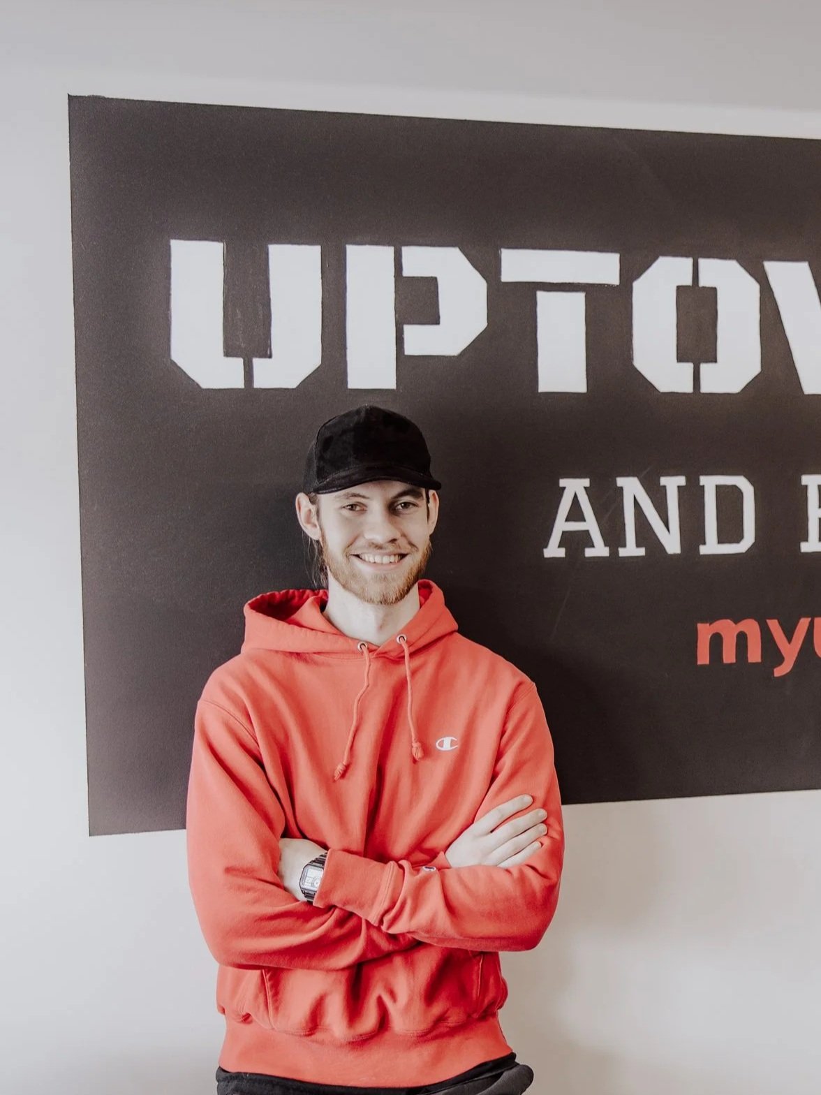 A young man with long hair, beard, and wearing a black cap and red hoodie, smiling and crossing his arms, standing in front of a black sign with white and red text.